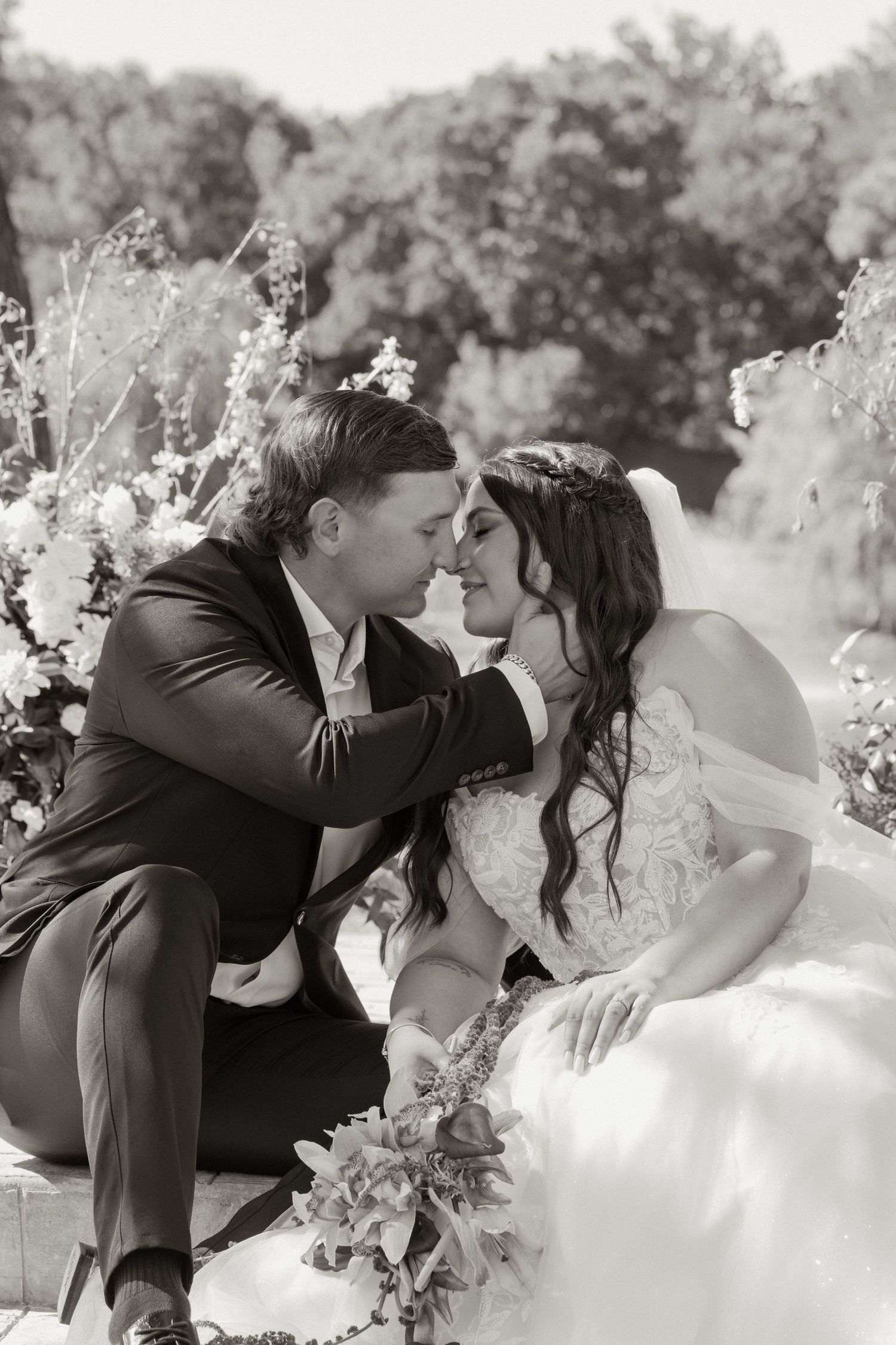 Black and white photo of a bride and groom sharing a romantic moment, seated among floral arrangements outdoors.