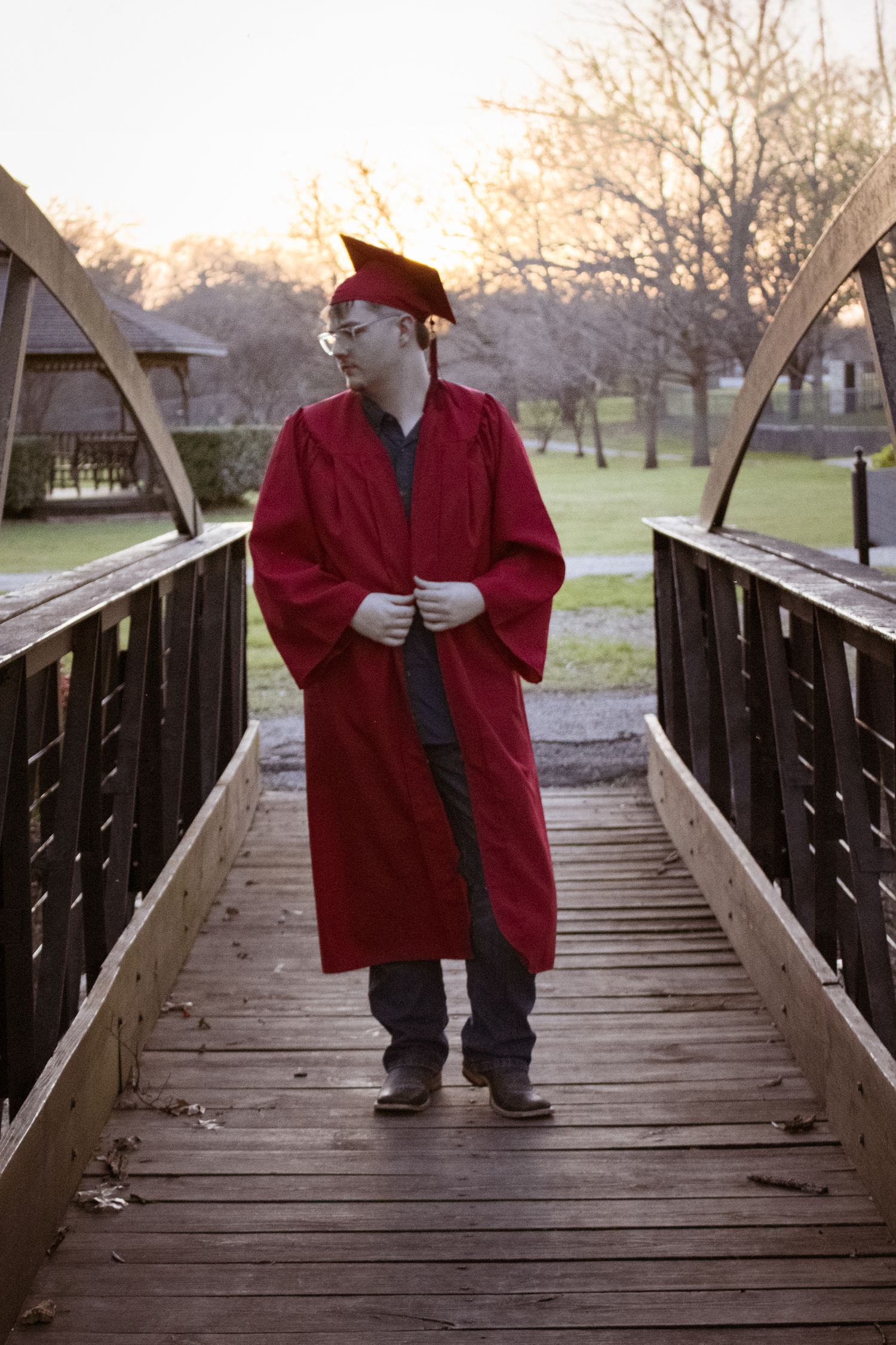Graduate in red cap and gown stands contemplatively on a wooden footbridge at sunset.