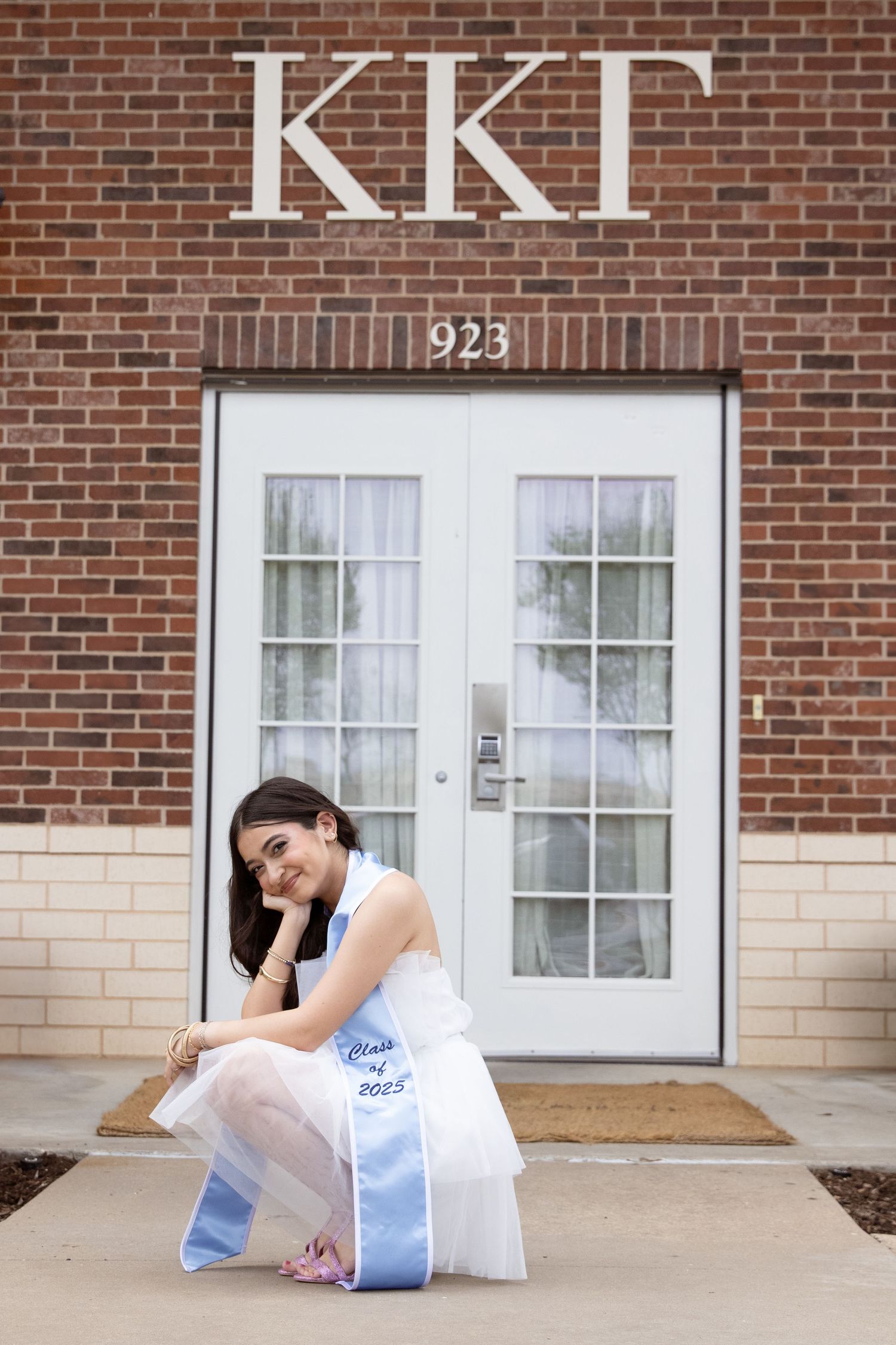 Graduate in white dress sitting in front of brick sorority house with KKG letters.