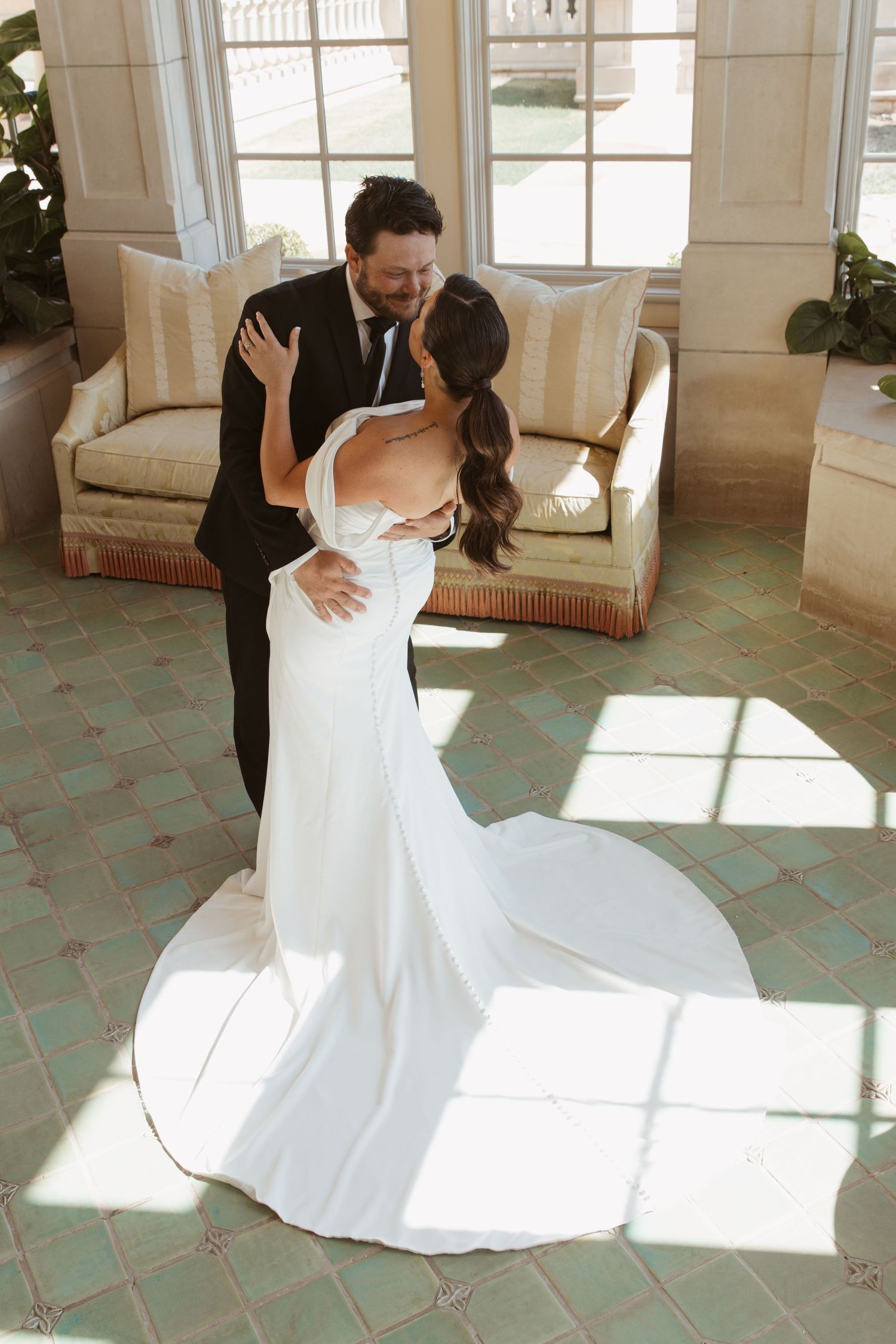 An elegant white wedding gown with train captured in a sun-lit interior space with tiled floors.