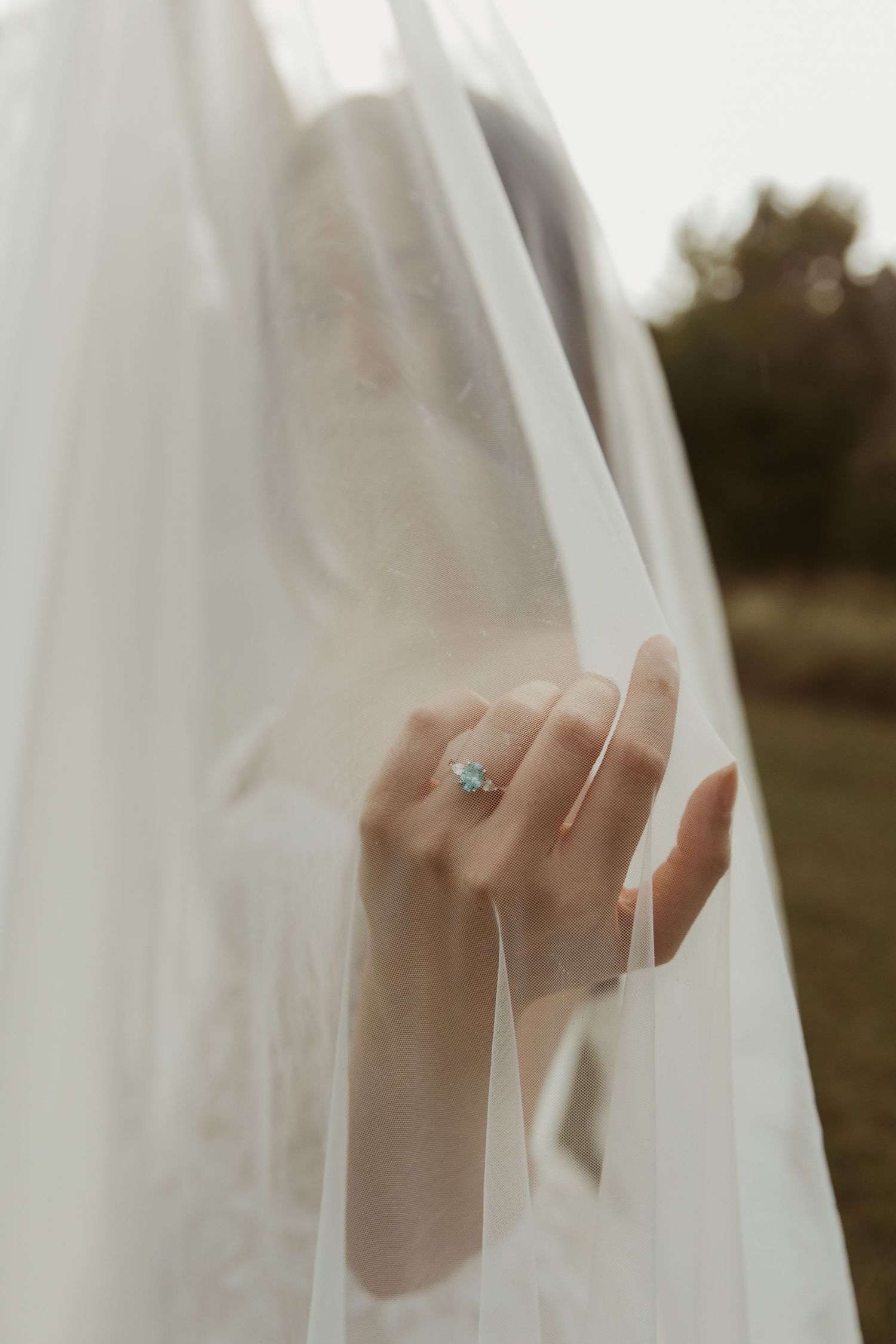 A delicate hand gently holds a sheer white veil against a soft, blurred outdoor background.
