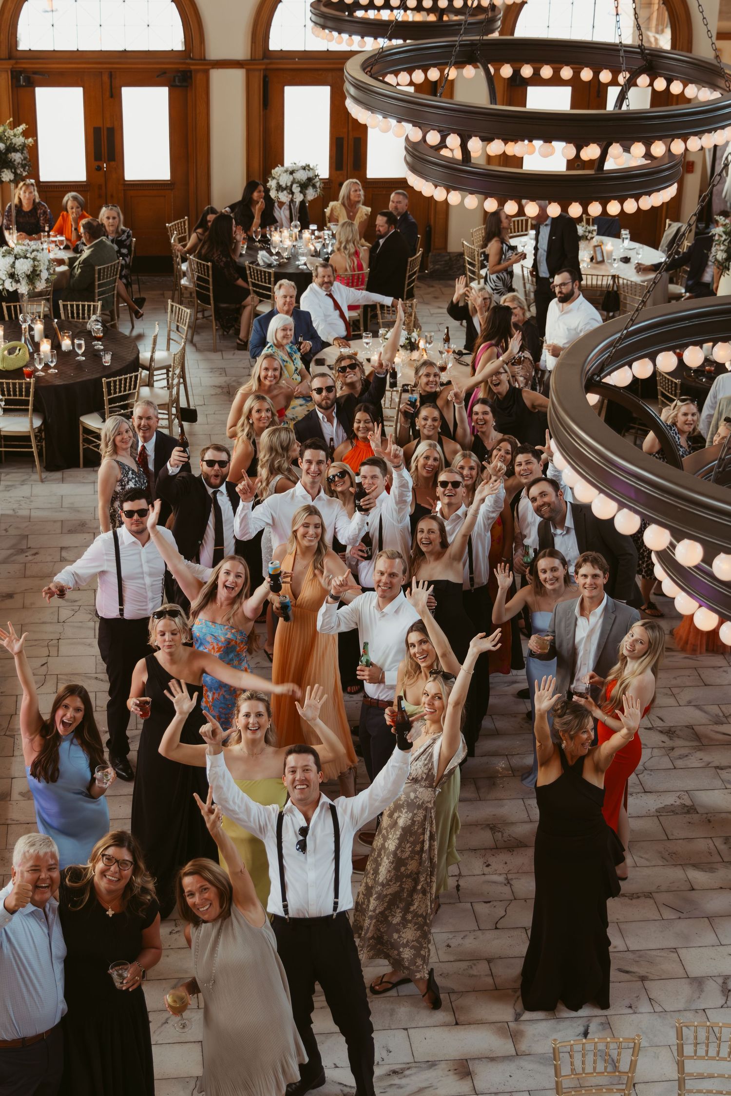 A large group celebrates at an elegant reception venue with tiered circular chandeliers and high arched windows.
