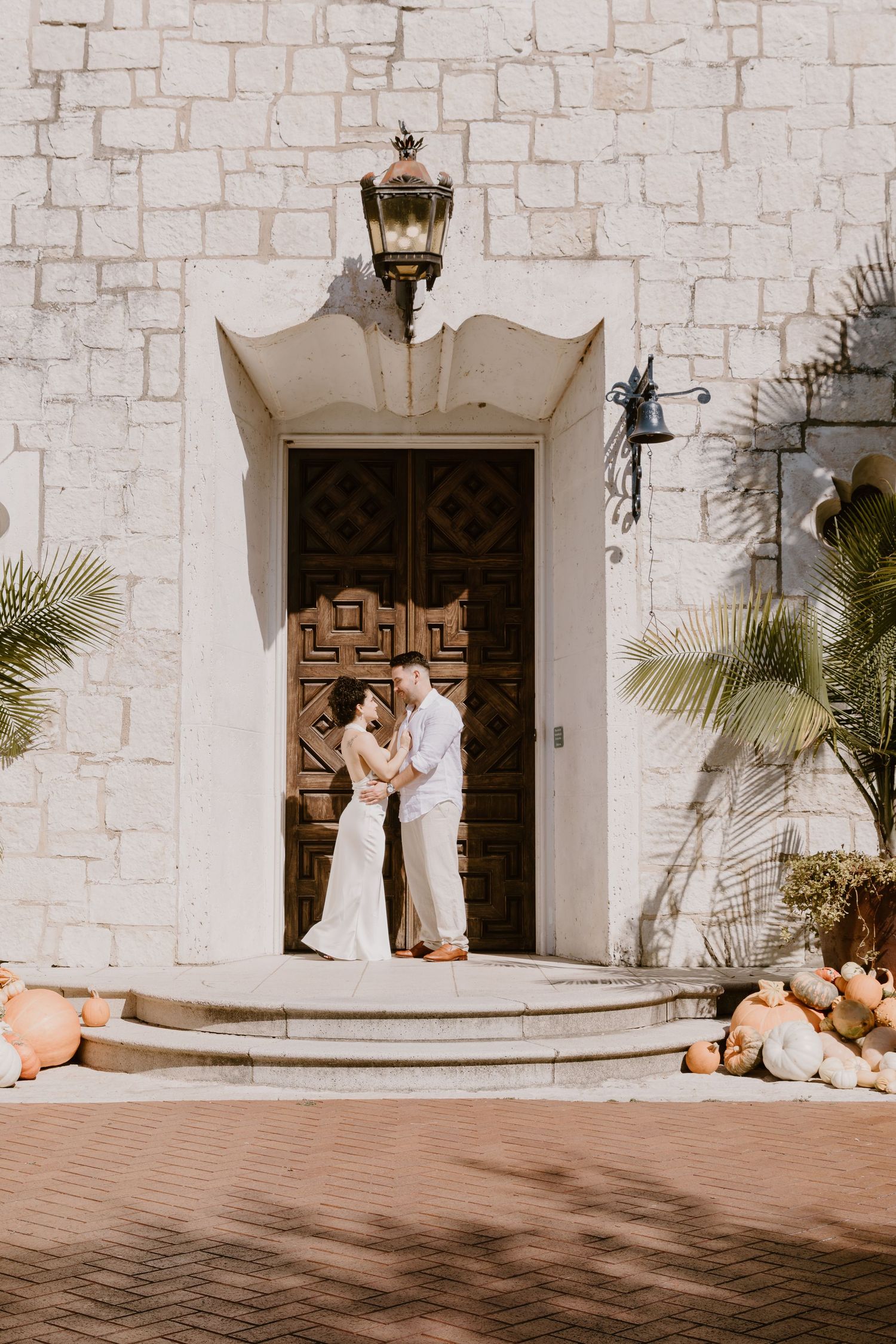 A couple shares tender moments in front of a white stone building with ornate wooden doors and palm trees.