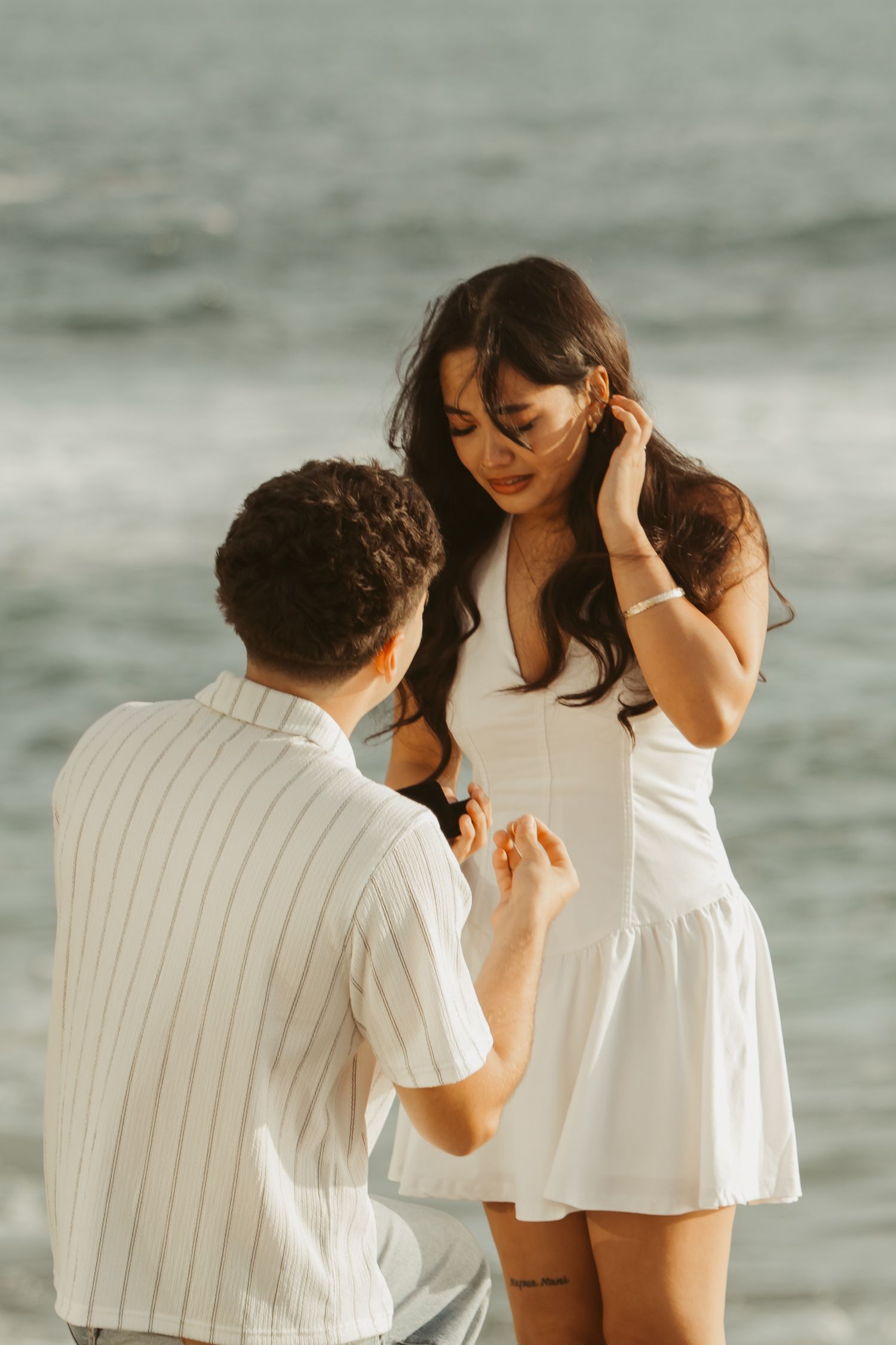 Man kneeling on beach proposing to woman in white dress near ocean.
