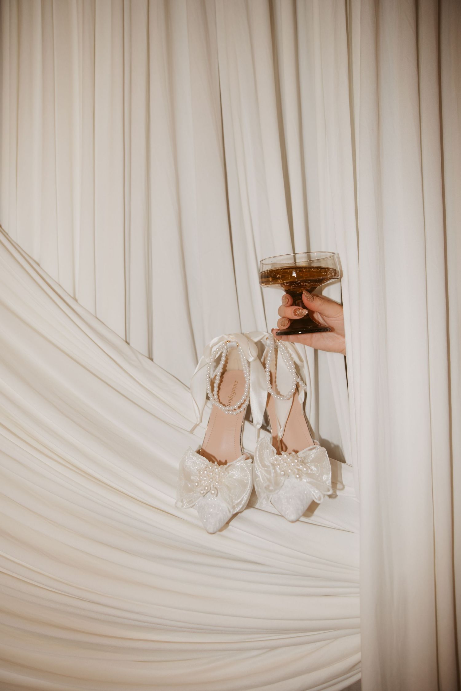Elegant white bridal heels with pearl embellishments displayed on draped ivory fabric with a glass candle holder.