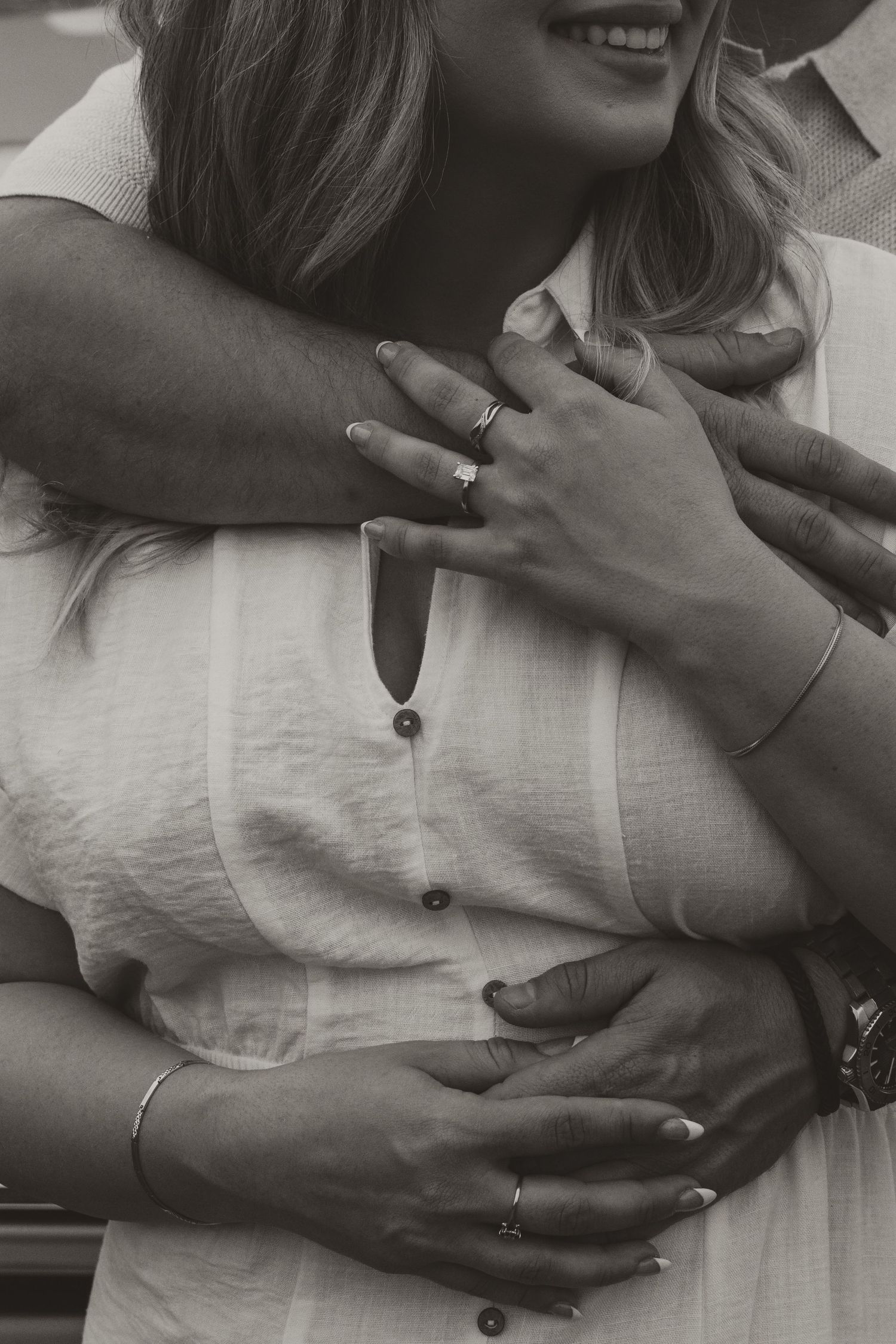 Black and white close-up of embracing couple wearing white clothing and jewelry showing hands with rings and bracelets.