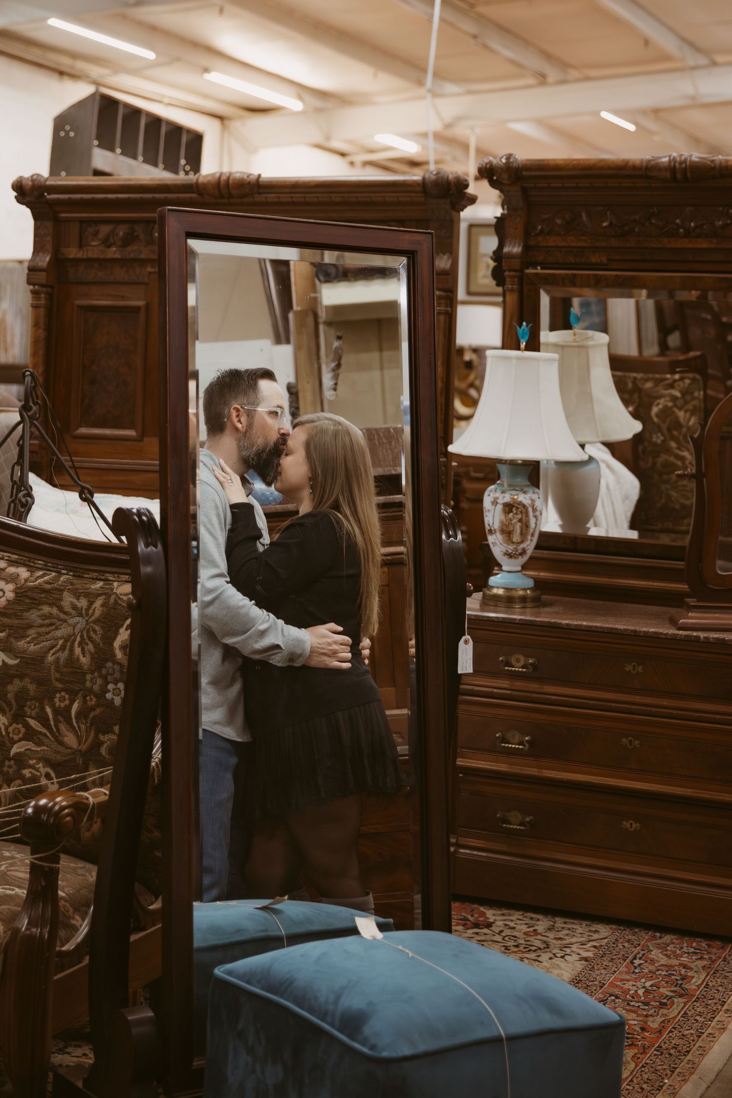 Couple browsing antique wooden furniture and decor in a vintage store with ornate mirrors and blue velvet ottoman.