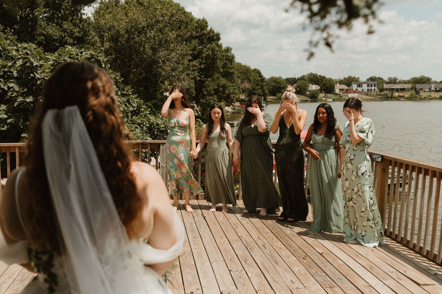 Wedding party in sage green dresses gathered on a waterfront deck sharing a joyful moment.
