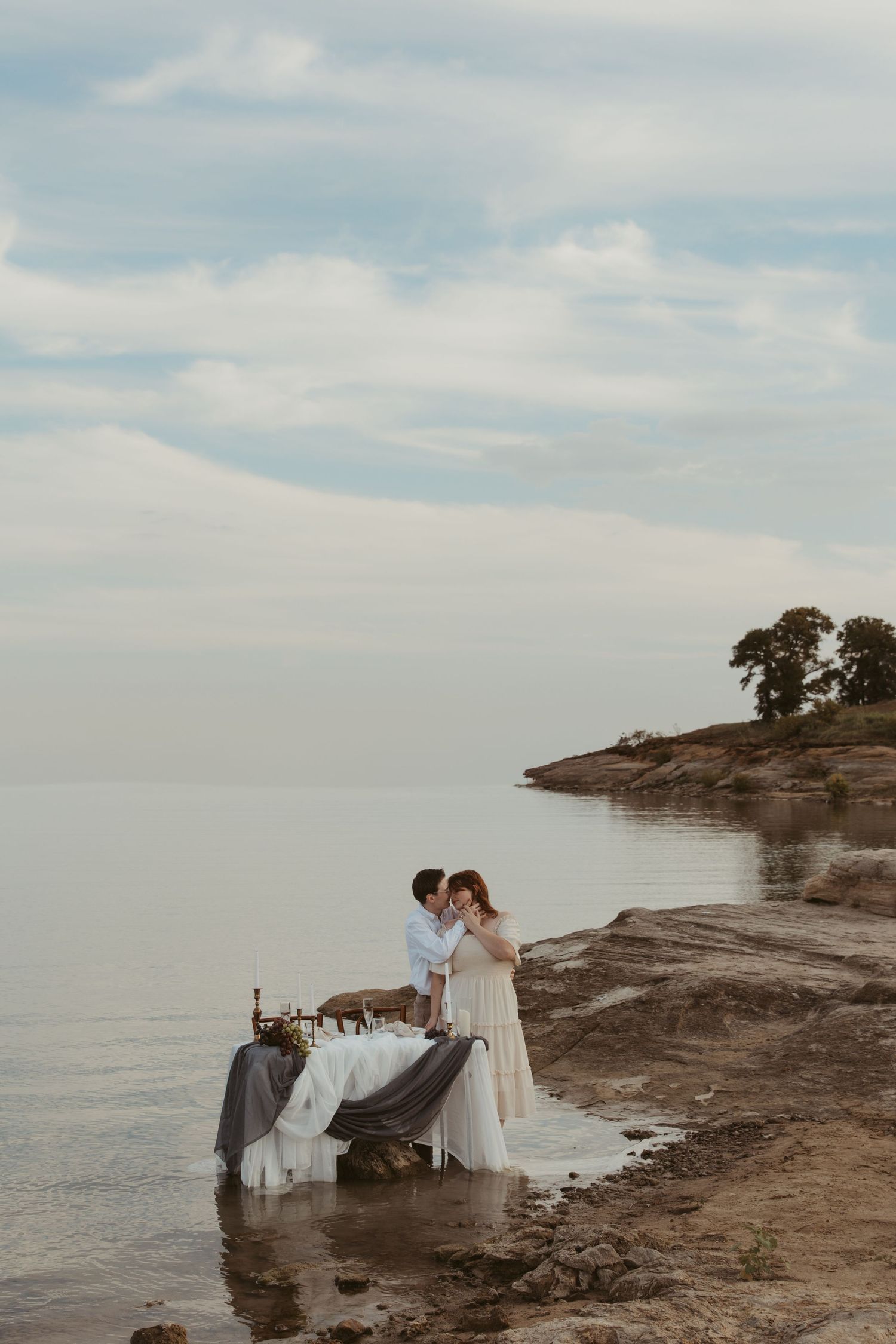 A romantic photoshoot takes place on a rocky coastal landscape with cloudy skies.