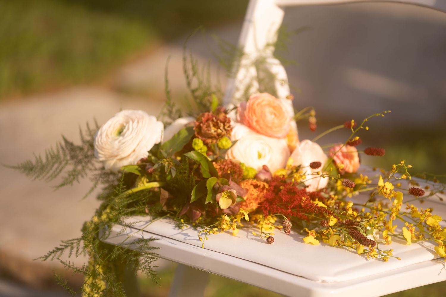 White chair adorned with a lush floral arrangement of peach roses, ranunculus, and yellow wildflowers at an outdoor wedding.