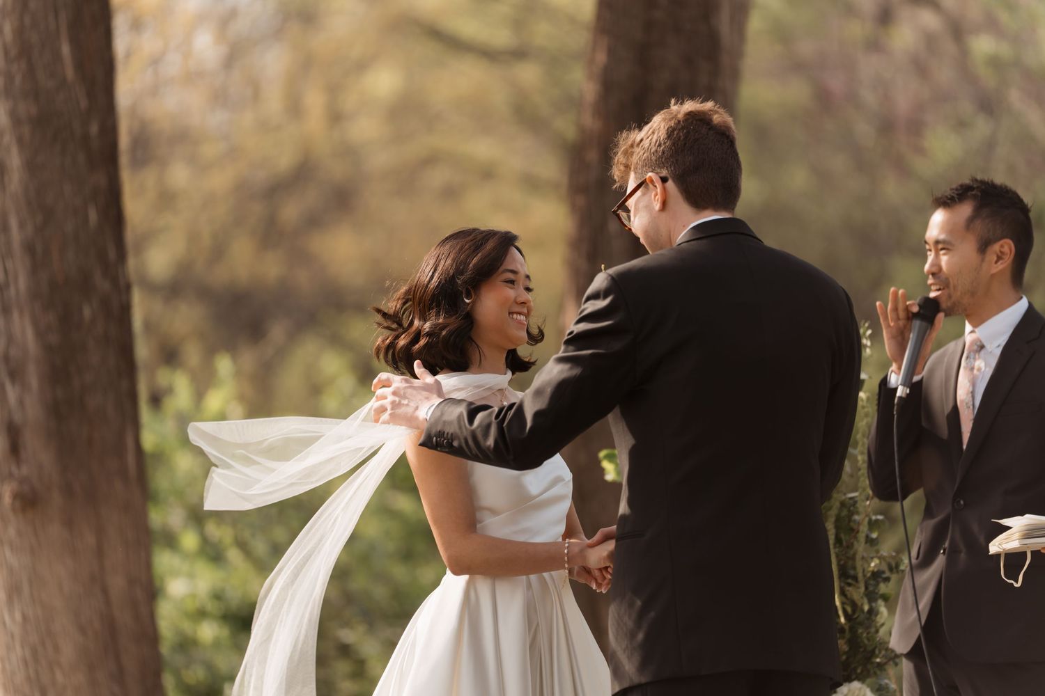 Bride and groom share a joyful moment during an outdoor wedding ceremony surrounded by trees.