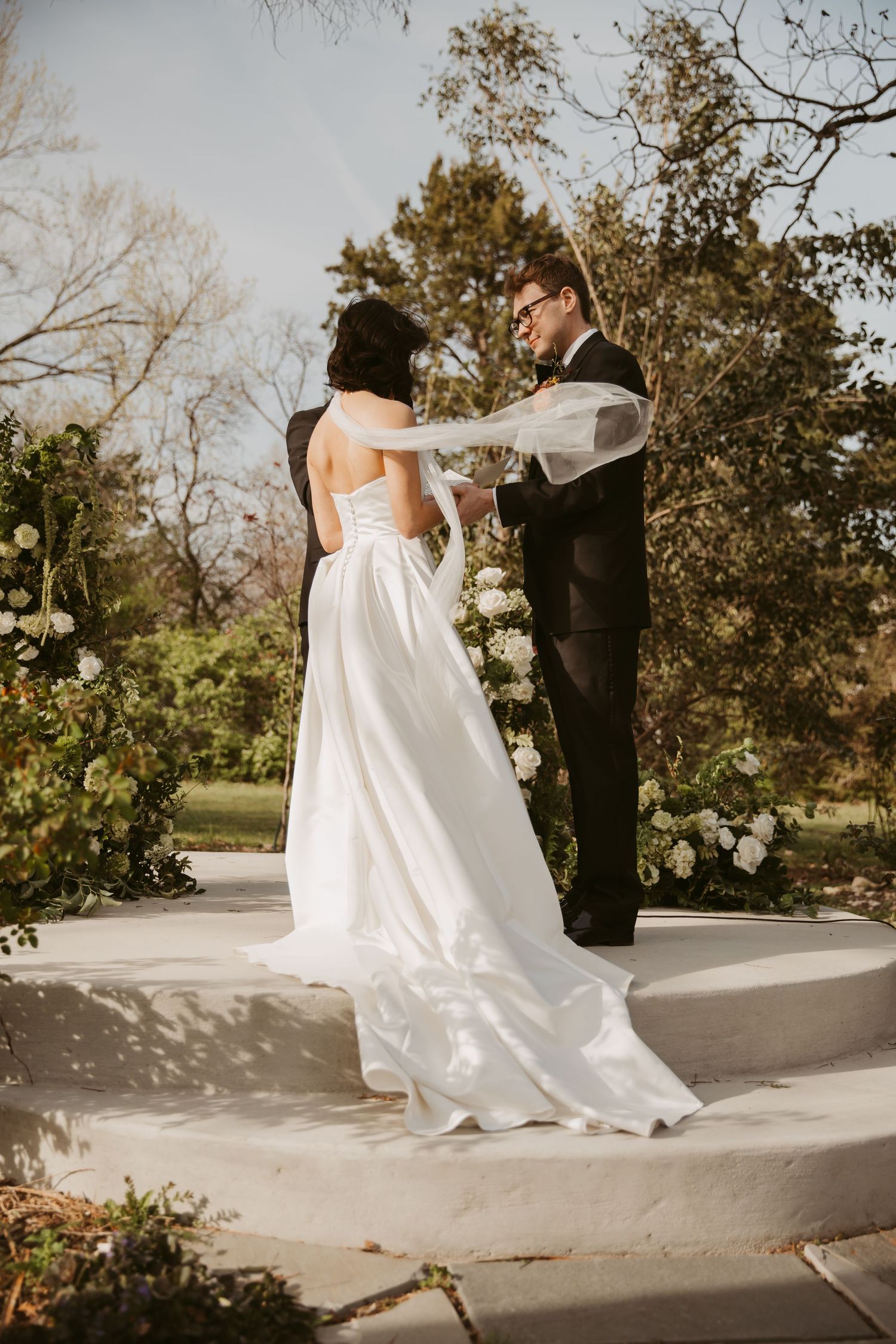 Groom holds brides veil during outdoor wedding ceremony on white circular platform with floral arch.