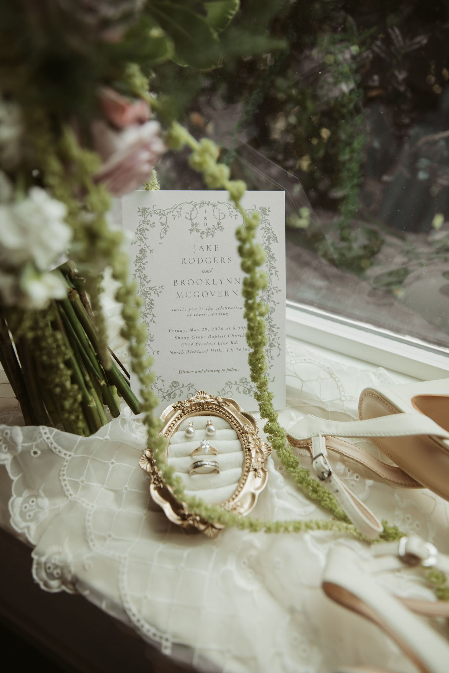 Wedding flat lay with invitation, rings, green floral necklace, and bridal shoes on lace fabric by a window.
