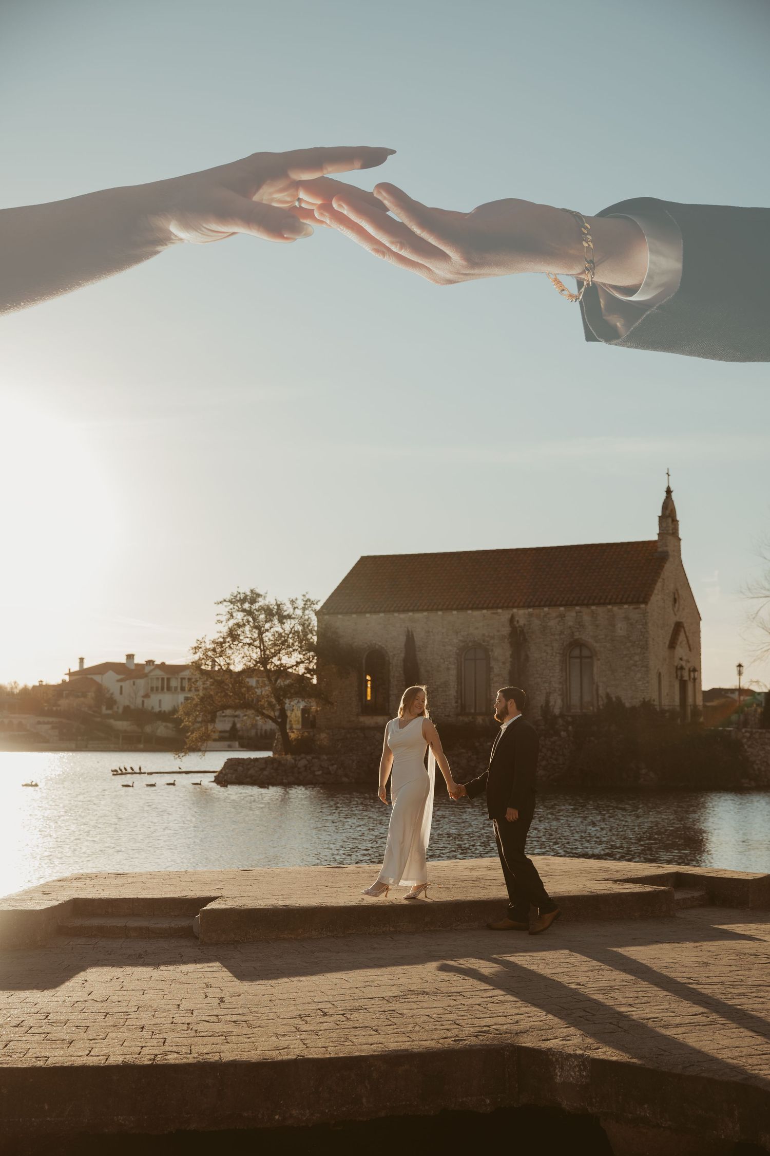 A couple poses on a stone pier at sunset with a historic seaside church and coastal landscape in the background.