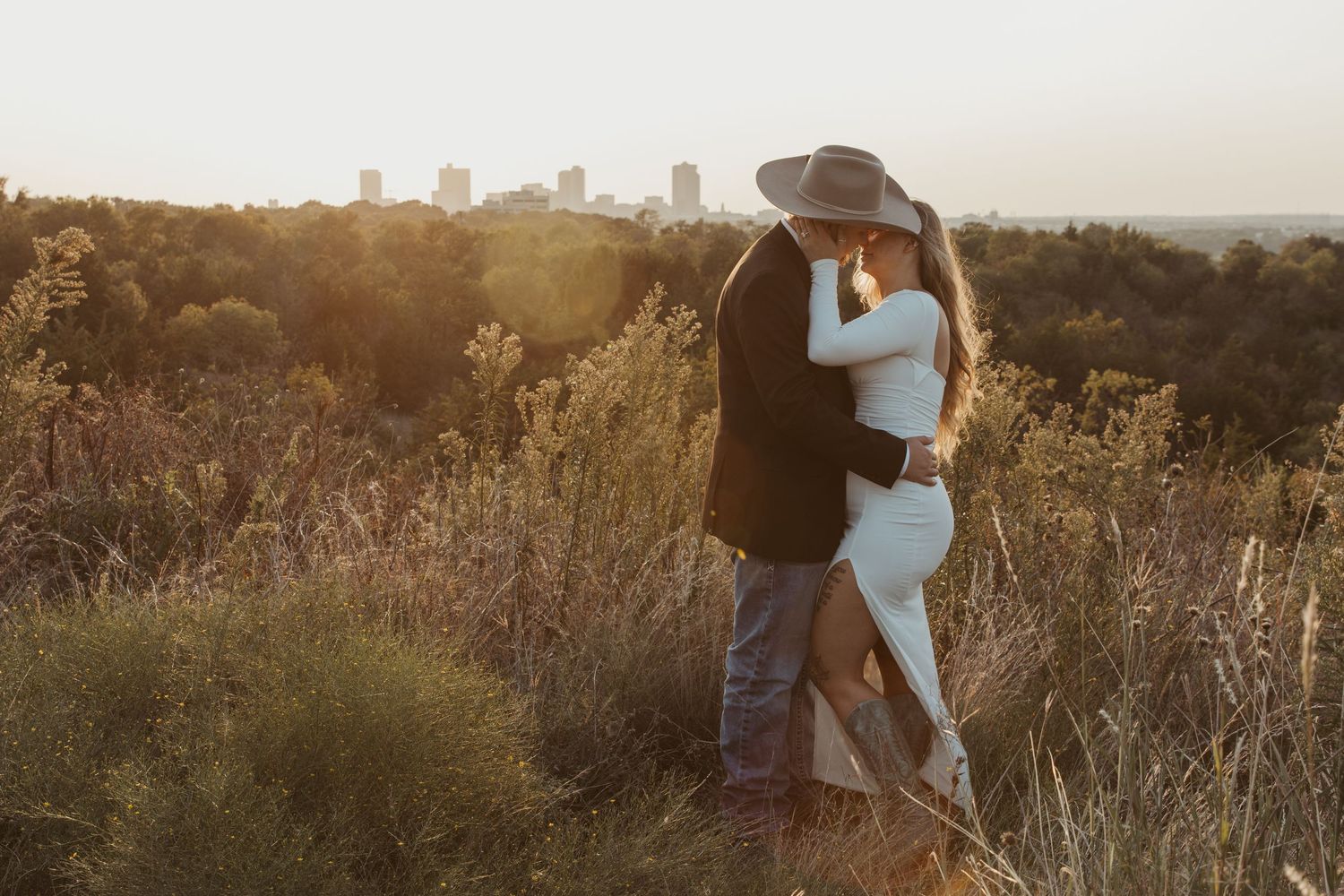 A couple embraces during a sunset photoshoot in a golden field with a cityscape backdrop in the distance.