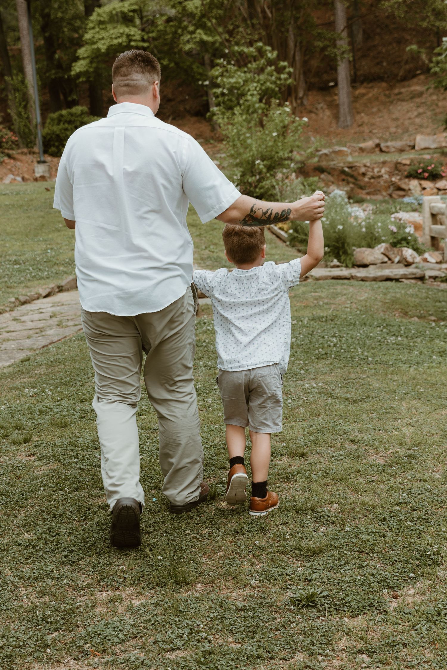 A parent and child walking hand in hand on grassy path, dressed in matching white shirts in a garden setting.