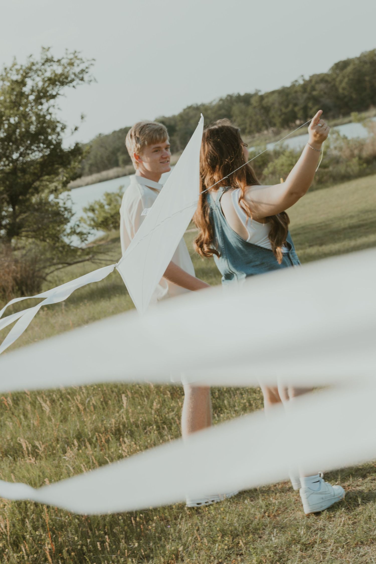 White flowing fabric creates artistic foreground blur while couple dances together on grassy hillside at sunset.