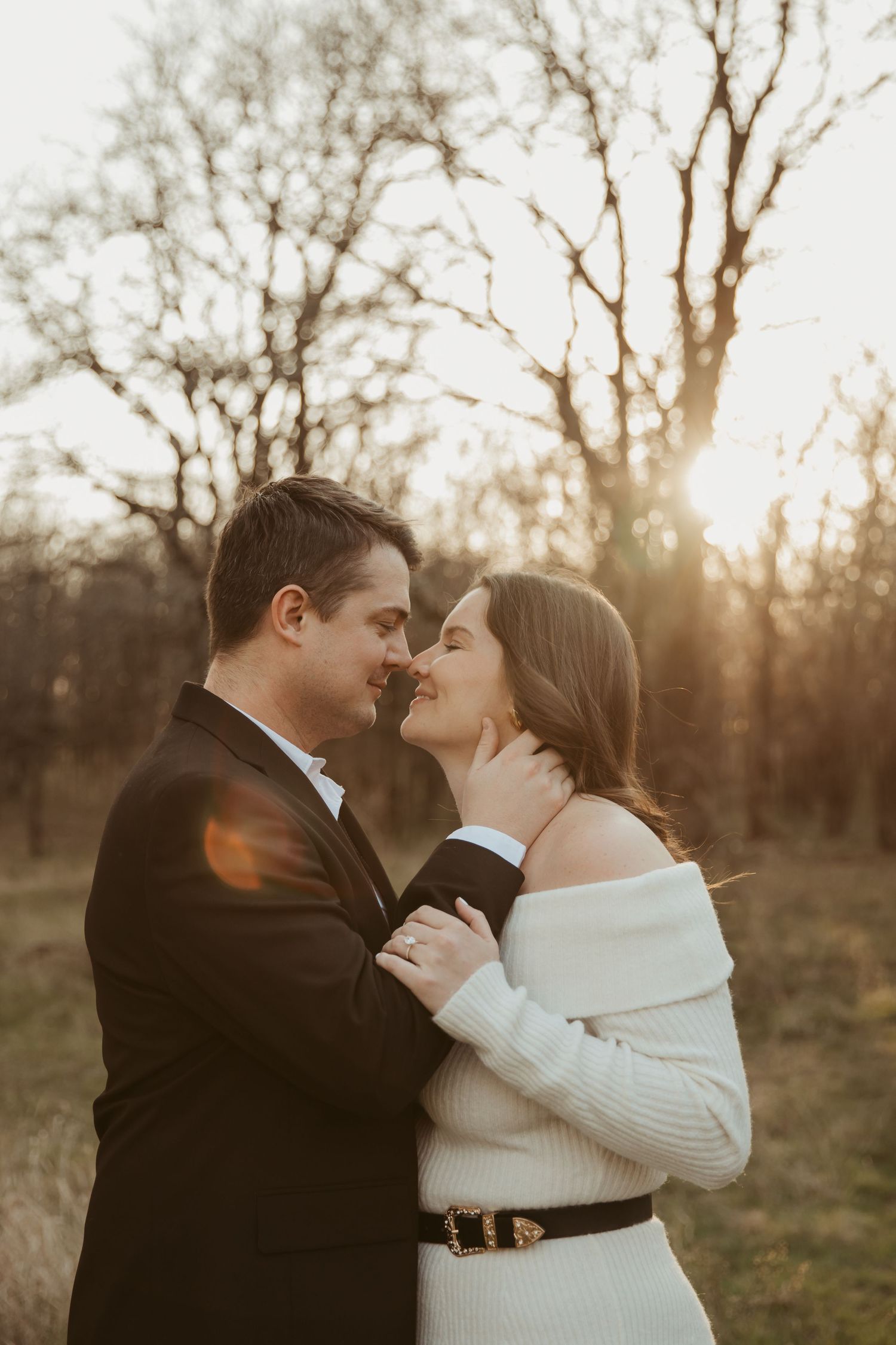 Couple sharing a romantic almost-kiss at golden hour, surrounded by bare trees in a sunlit outdoor setting.