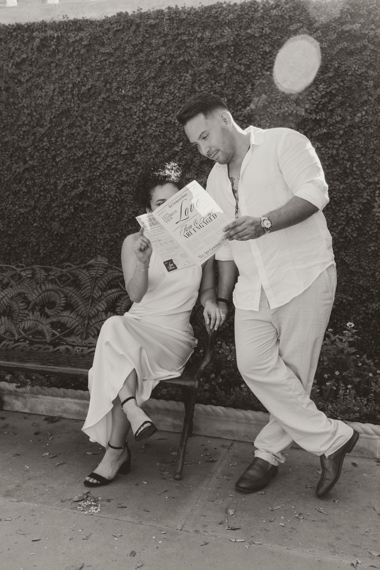 A couple in matching white outfits shares playful moments on a bench in black and white photography.