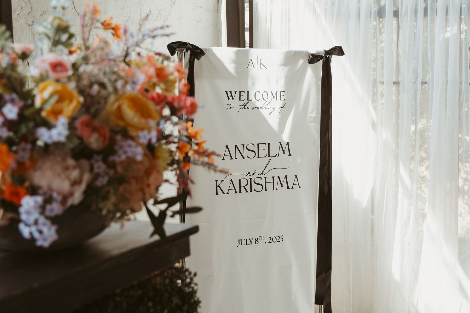 A wedding welcome sign with text displayed next to a floral arrangement near white curtains.