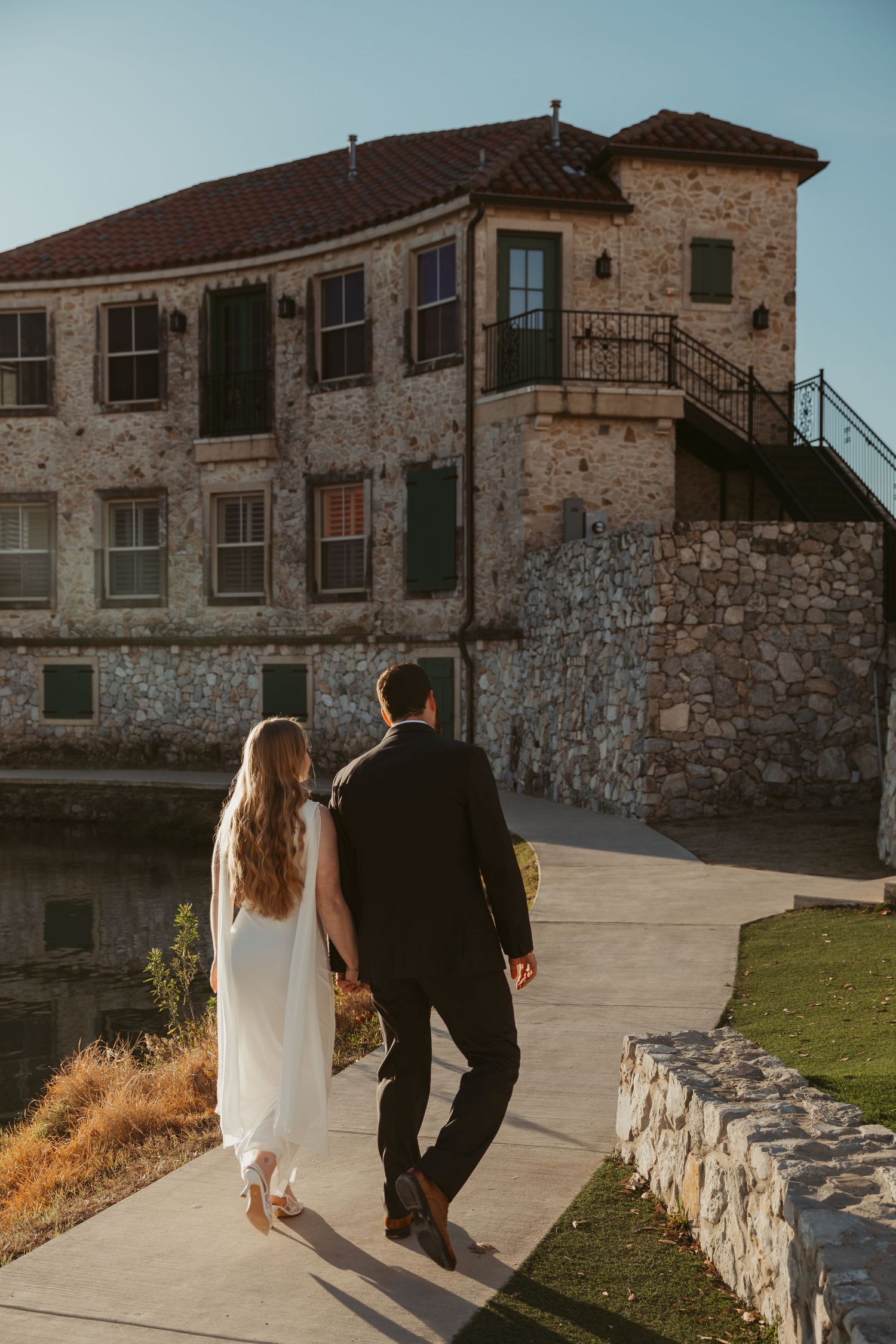 Couple walks hand in hand past historic stone buildings on their wedding day.