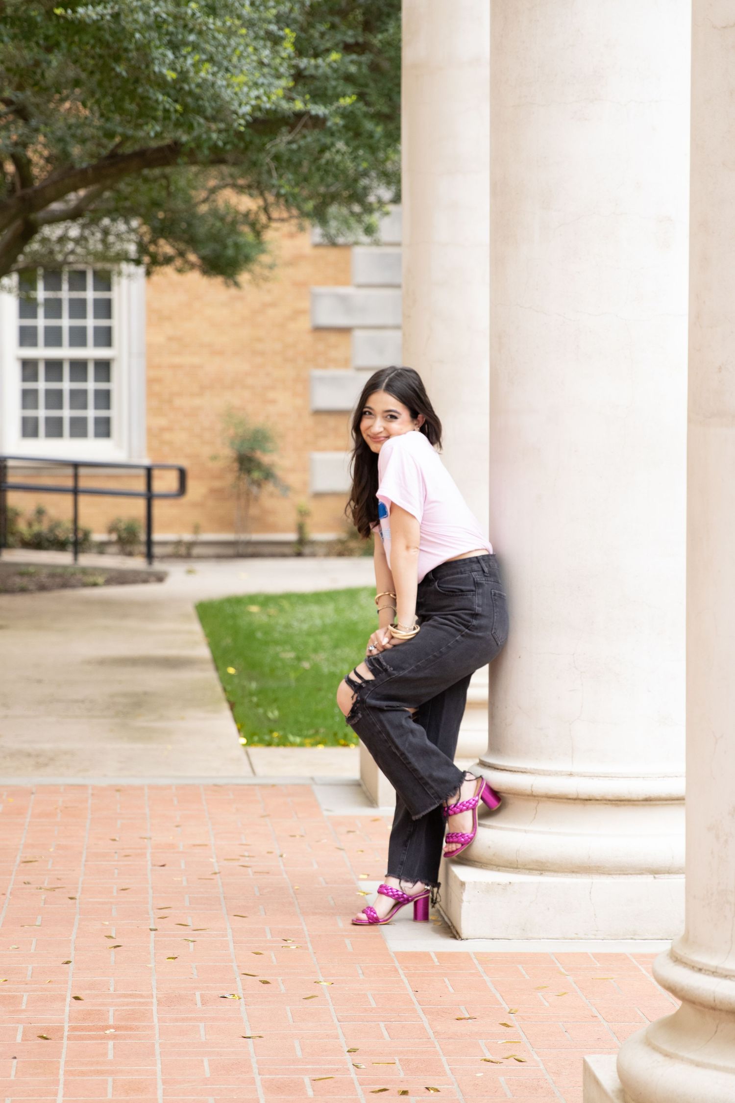 Someone poses playfully against a white column on a brick walkway wearing pink heels and dark pants on a college campus.