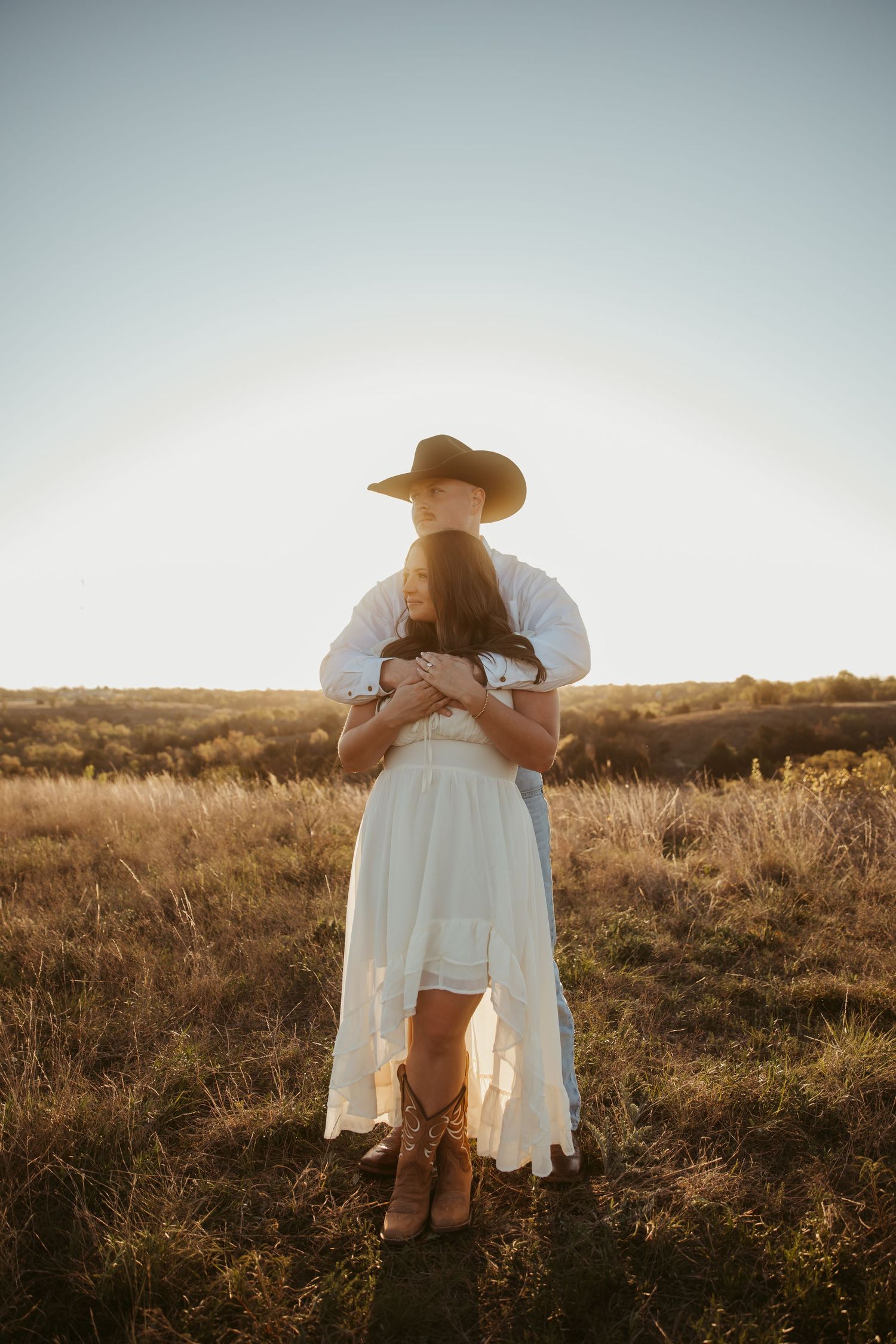 Couple embracing in golden field at sunset, woman in white dress and cowboy boots, man in cowboy hat and white shirt.