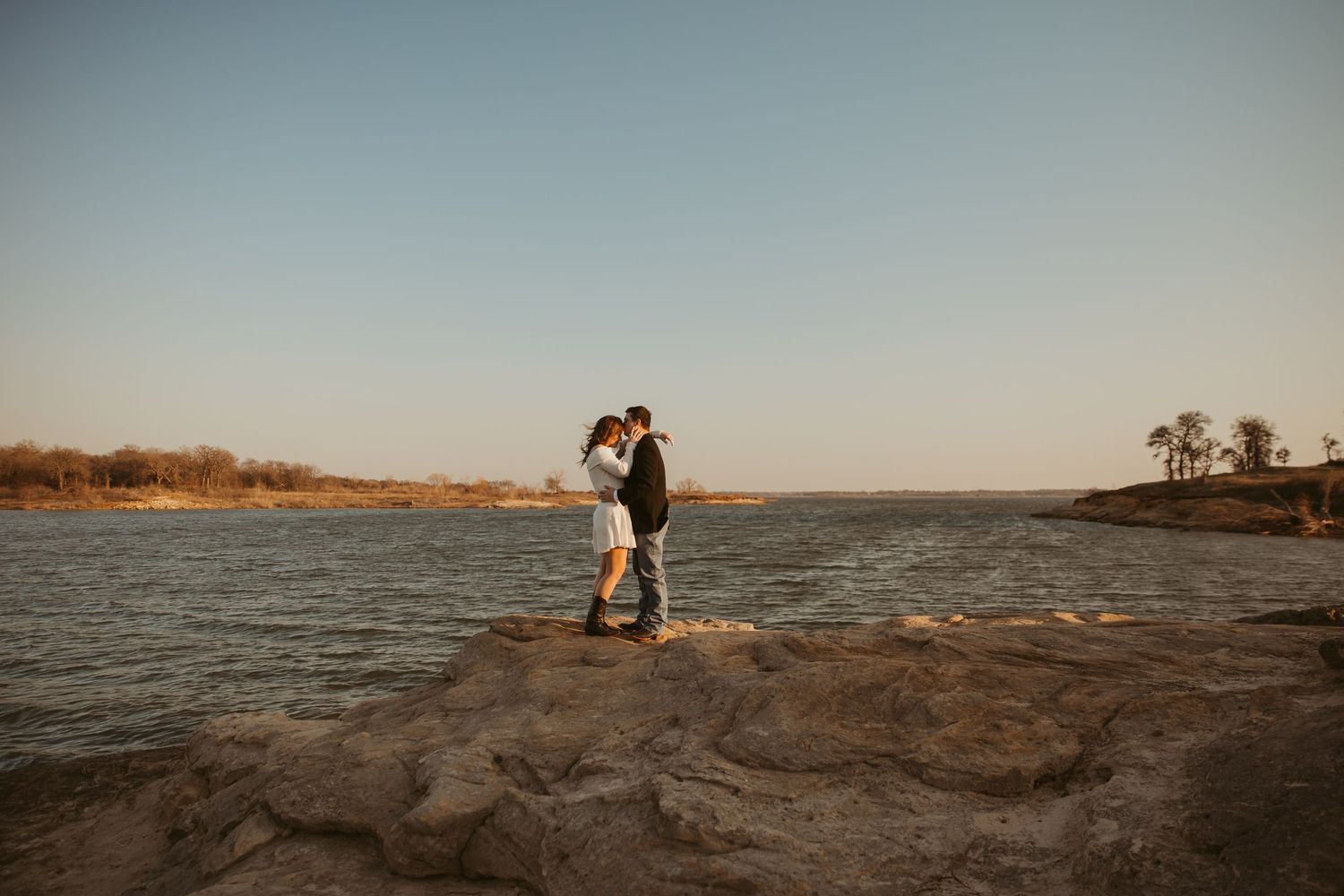 Couple embracing and kissing on rocky shoreline at golden hour with calm river and trees in background.
