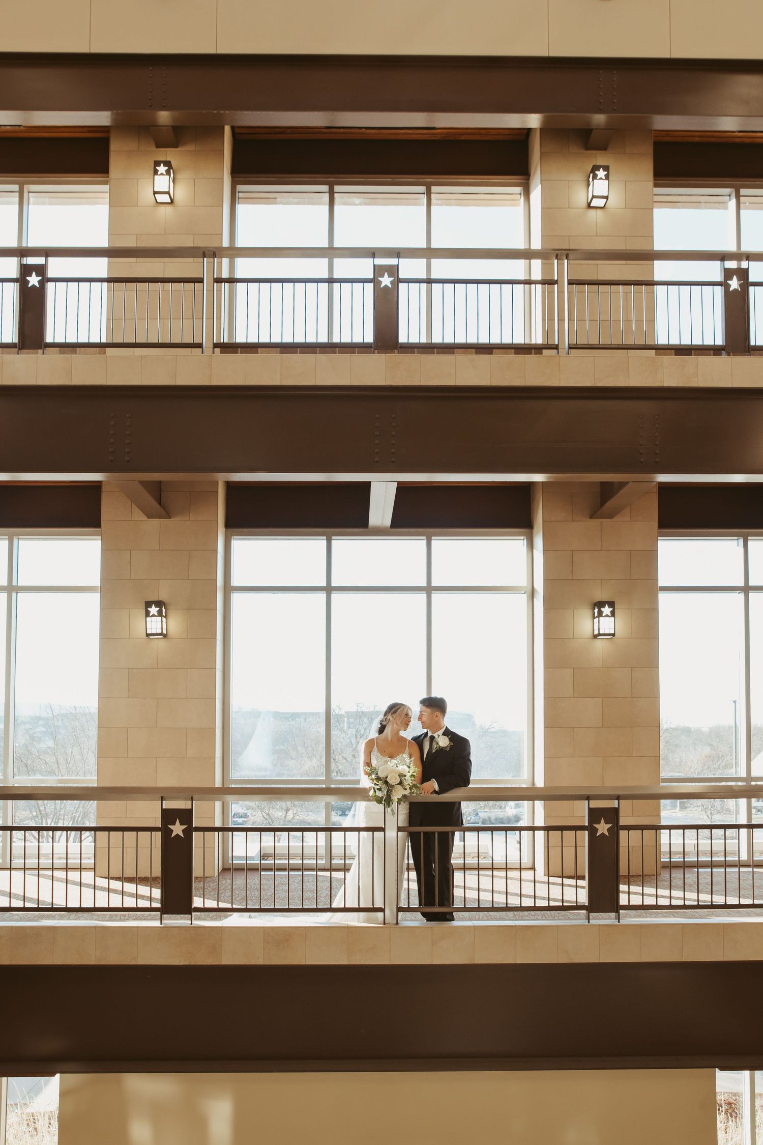 A couple shares a tender moment on a balcony overlooking large windows in an elegant indoor venue.