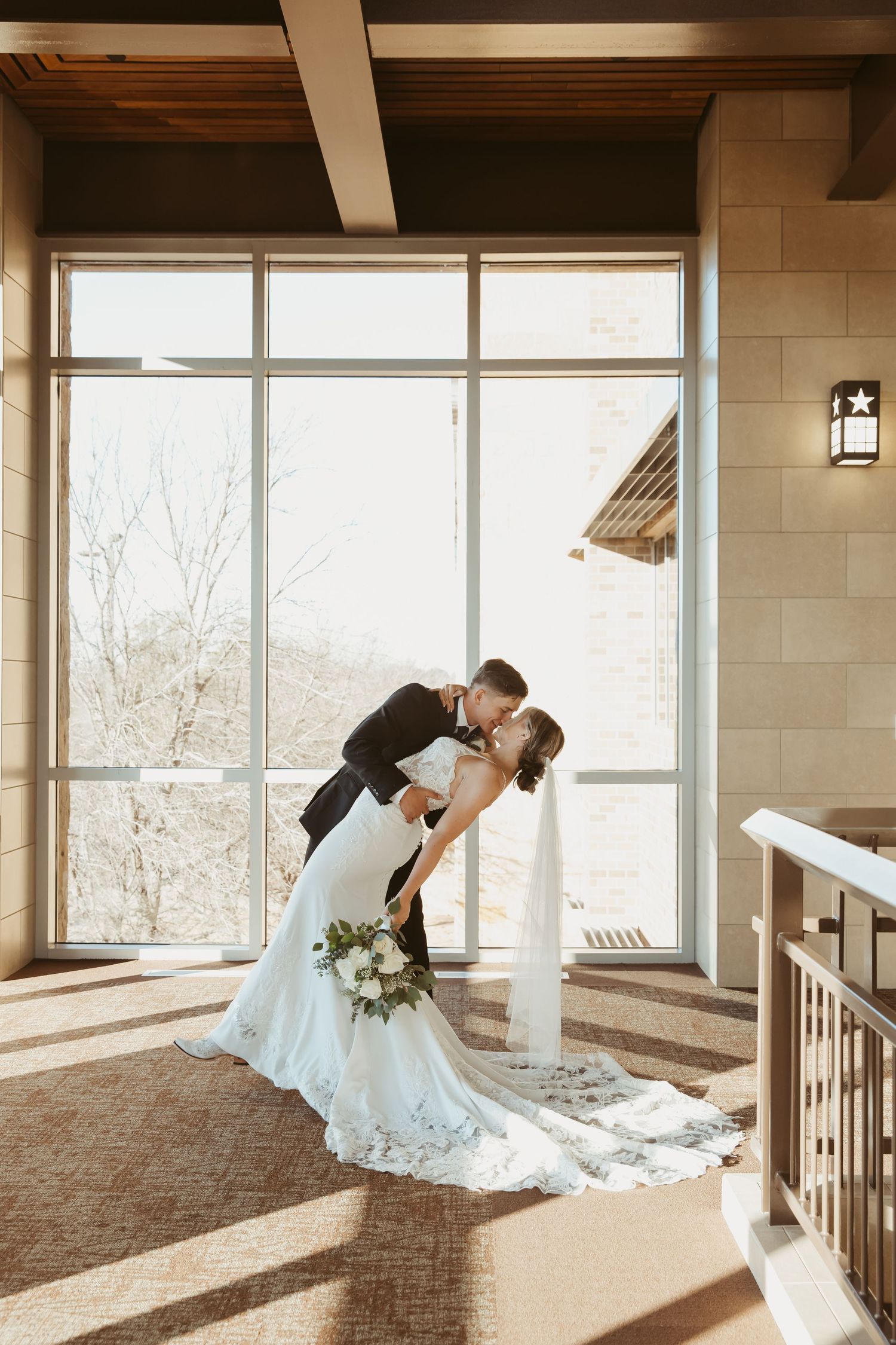 A romantic couple shares an intimate dip pose in front of large windows in an elegant indoor venue.