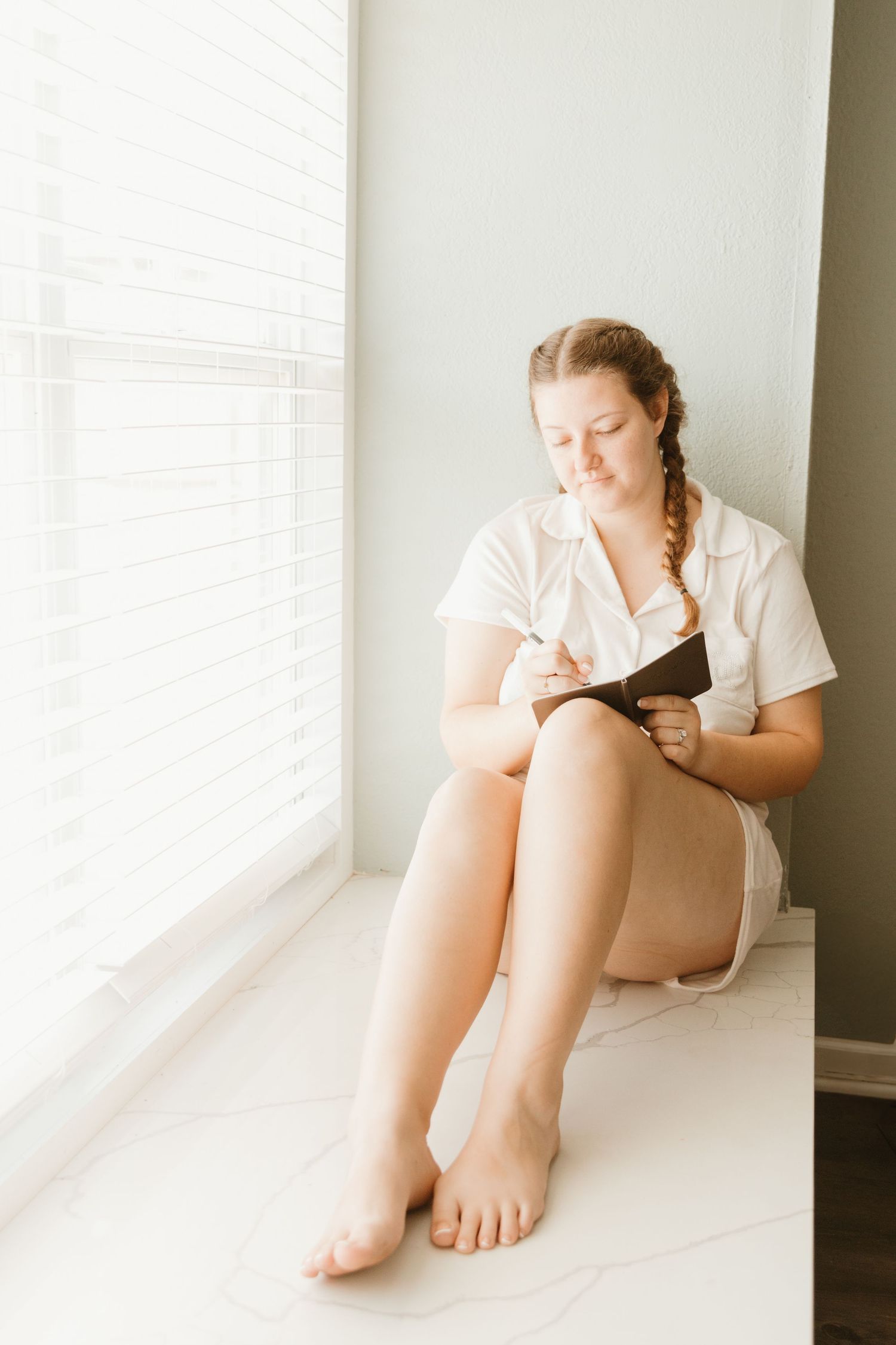 A person in white clothing sits by a window writing in a journal on a sunny morning.