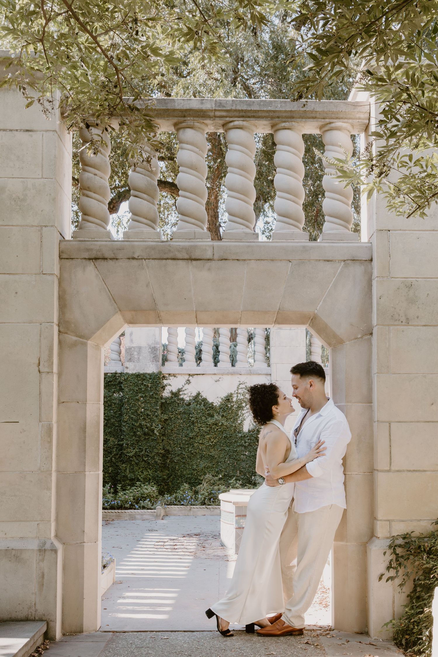 A couple in white outfits sits together on a stone bench under an elegant colonnade with hanging vines.