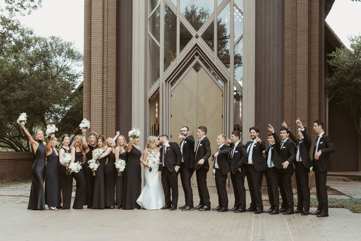 Elegant wedding party stands in traditional formation against stone church facade in courtyard during formal photo session.