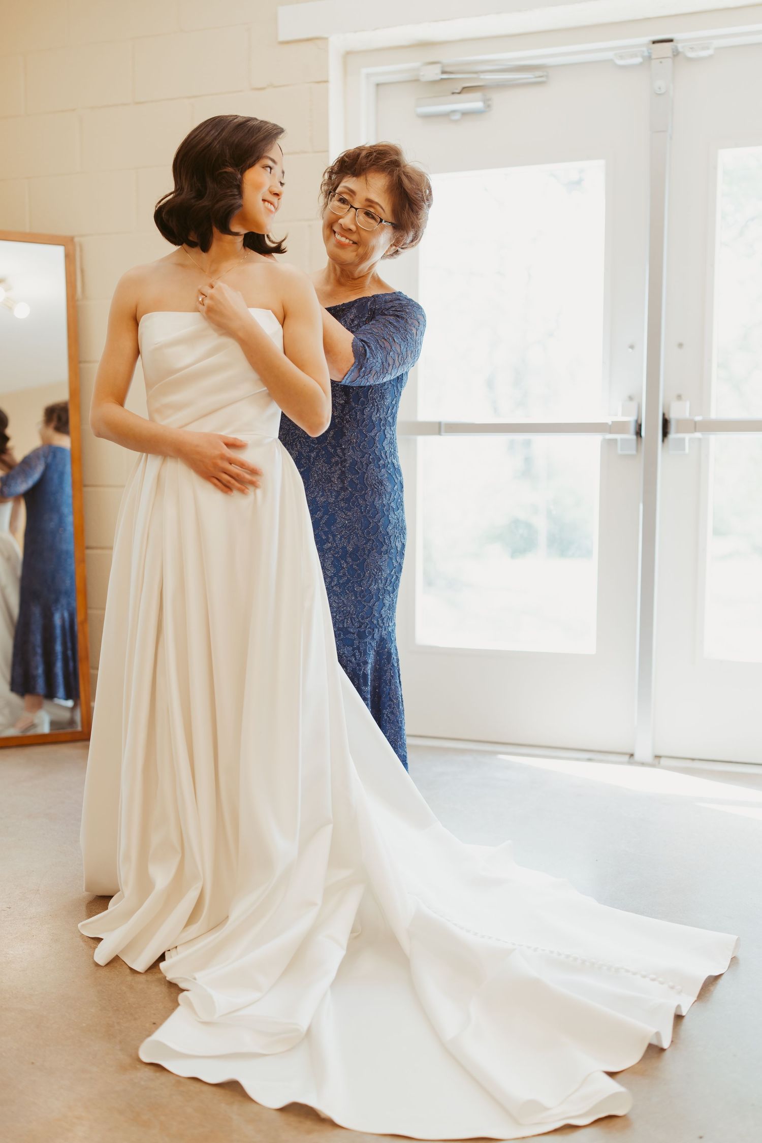 Bride in white strapless gown being helped by older woman in blue lace dress near bright window on wedding day.