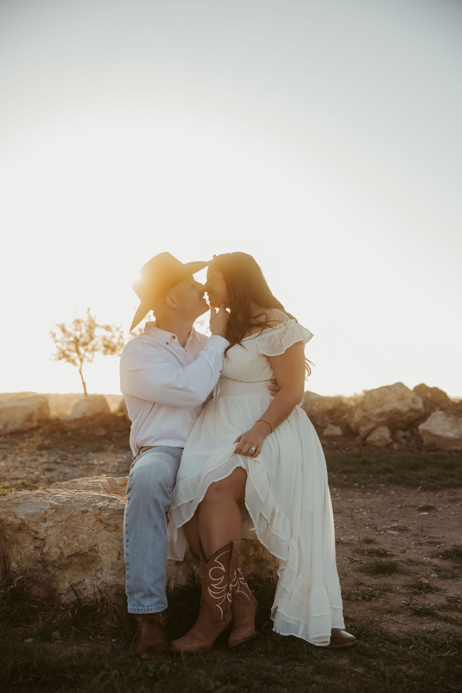 Couple sharing a romantic moment at sunset, man in cowboy hat and jeans, woman in white boho dress and cowboy boots.