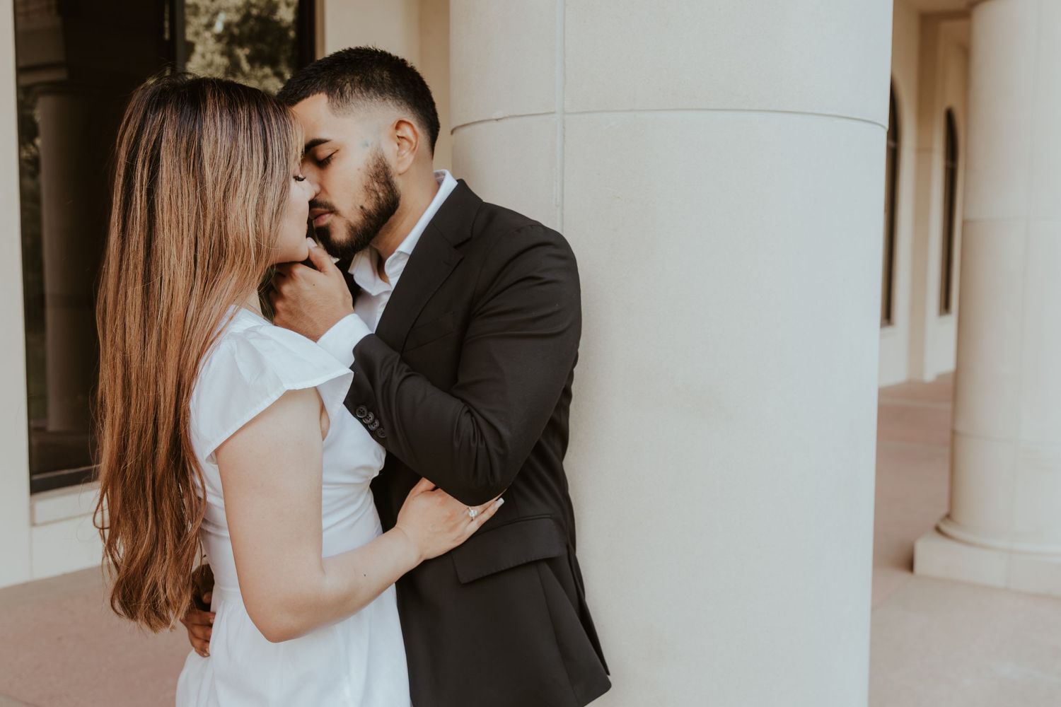 A couple wearing face masks embrace in a romantic moment between white columns in an elegant indoor setting. Engagement photographer DFW