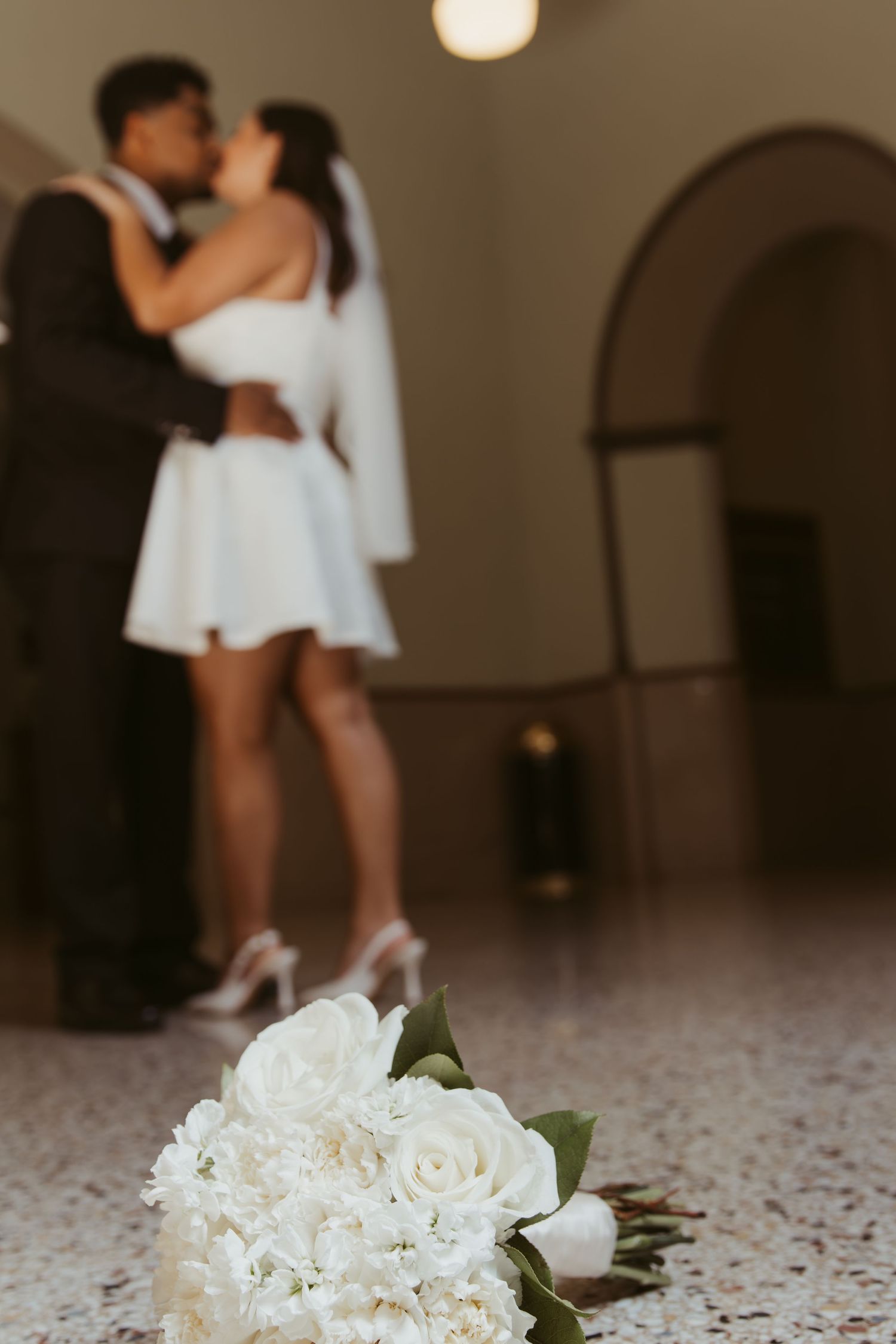 A white floral bouquet rests on a speckled floor with a couple dancing in soft focus above. Wedding Photographer DFW