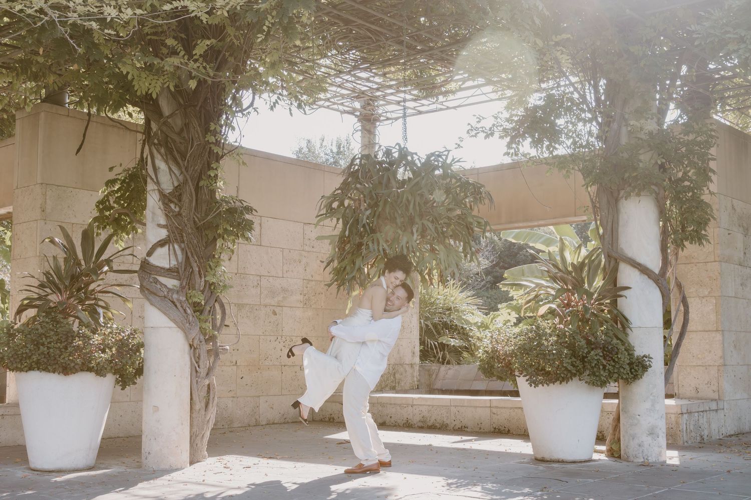 A person in white clothing dances on a sunlit patio surrounded by potted plants and climbing vines.