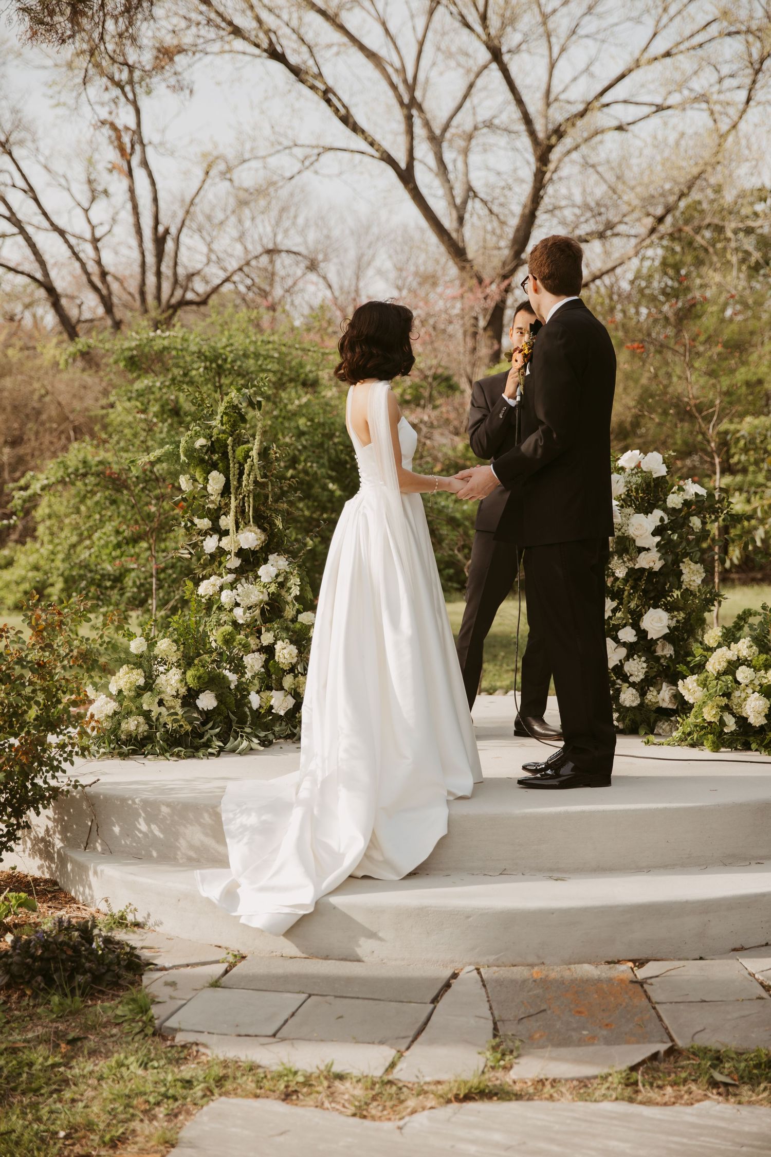 Bride in white gown and groom in black tuxedo exchanging vows at outdoor garden ceremony.