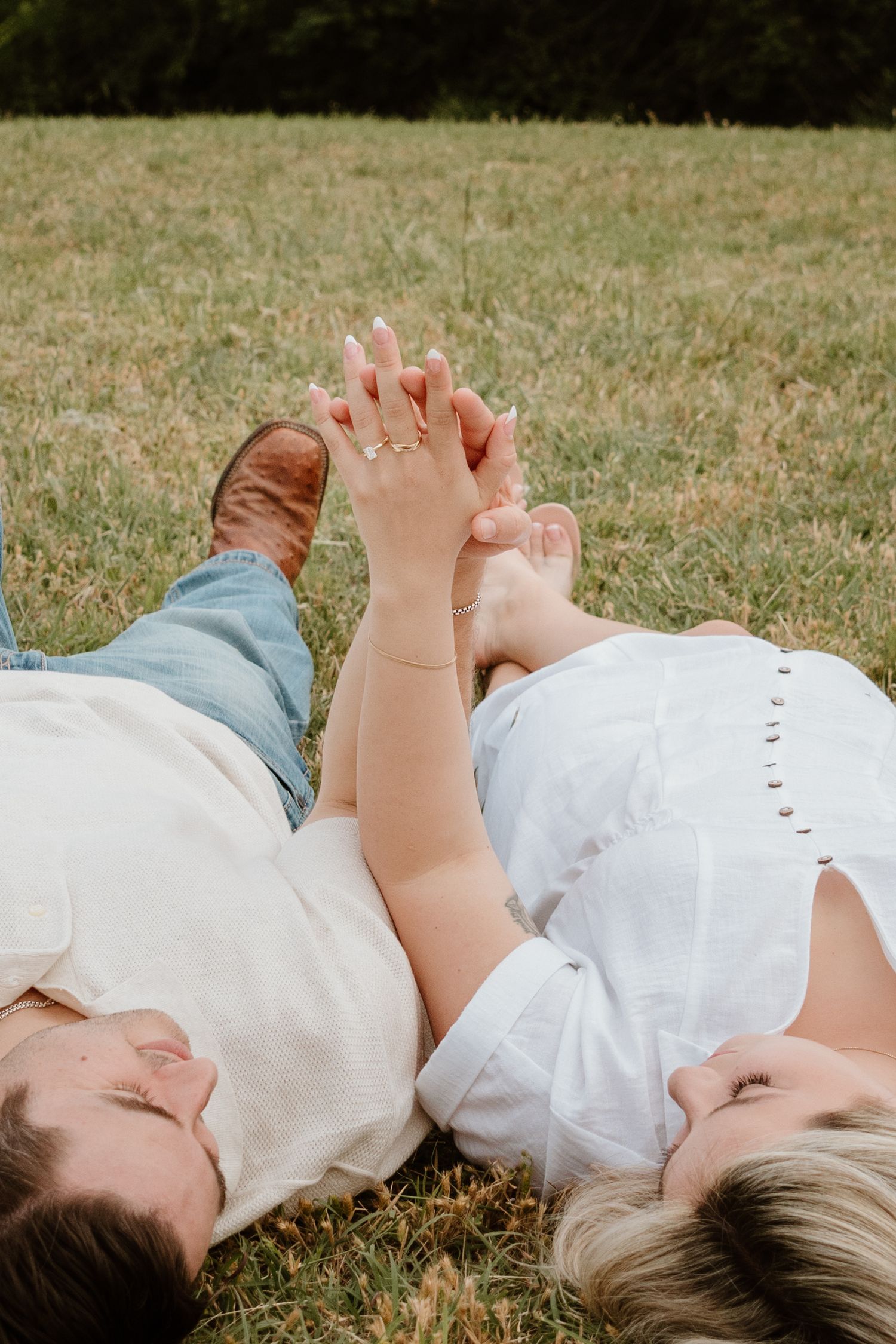 Two people lying on grass with their feet touching while wearing casual clothing in a sunny outdoor setting.