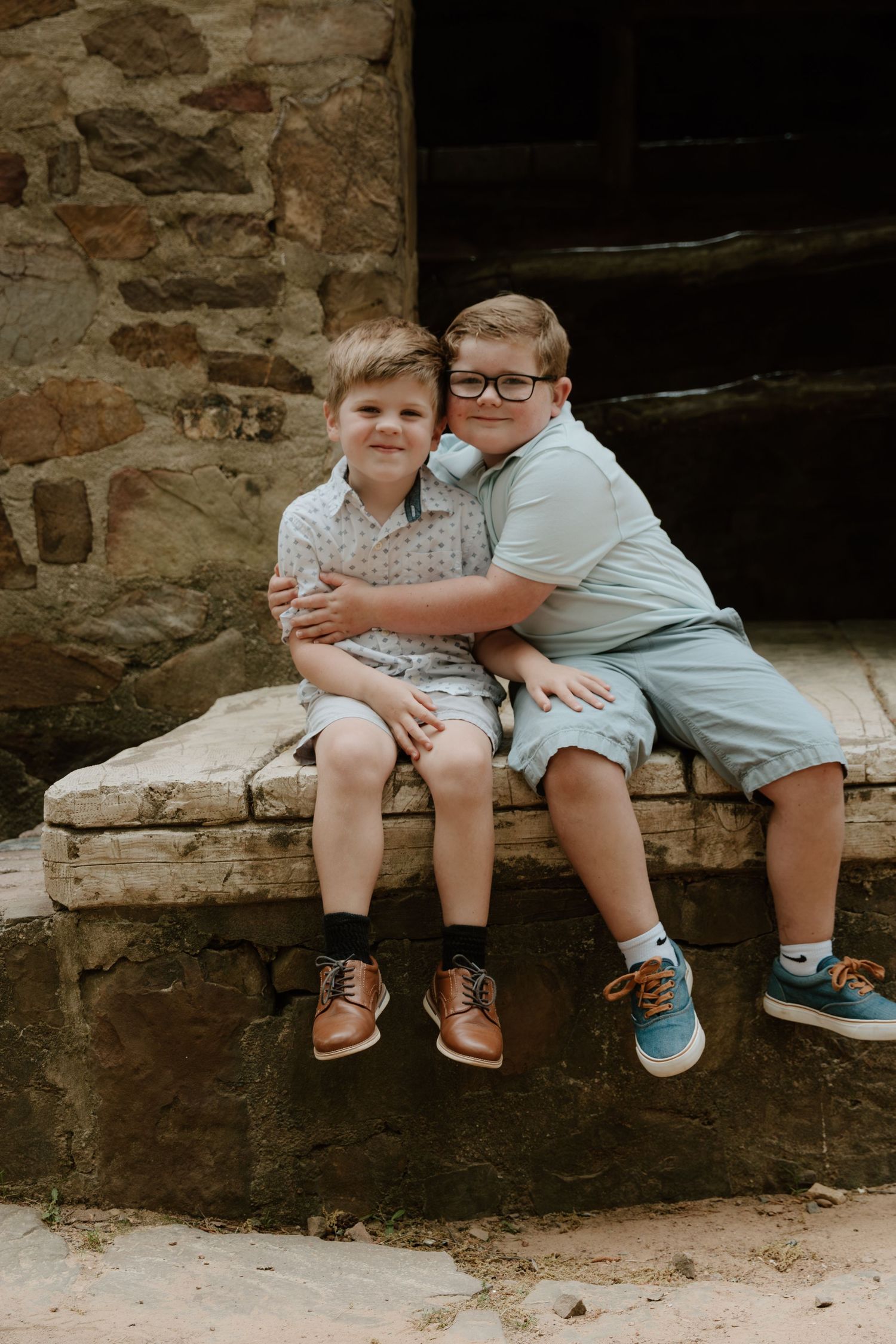 Two young children sit together on stone steps outside a rustic building wearing casual summer clothes and sneakers.