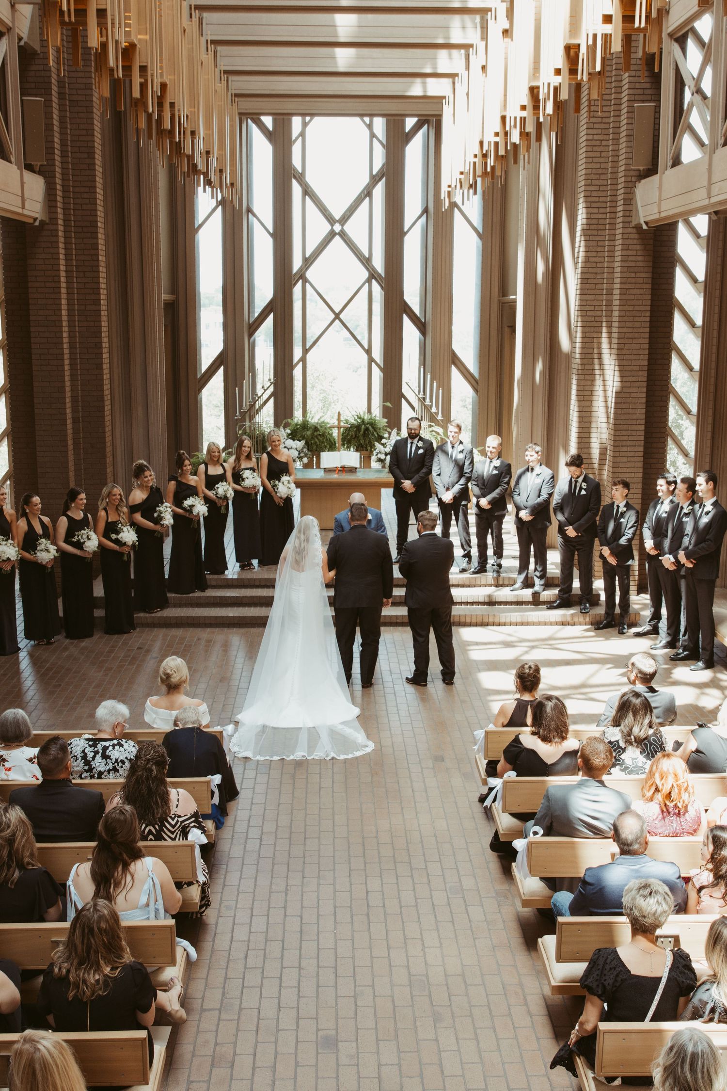 Wedding ceremony taking place in a modern chapel with high wooden beams and guests seated in wooden pews along the aisle.