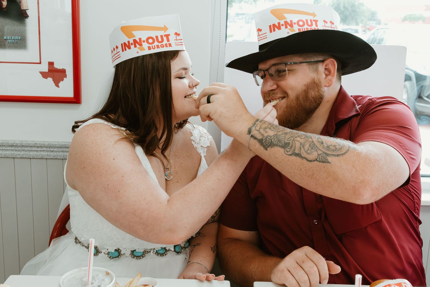 Newlyweds wearing In-N-Out paper hats share a lighthearted moment feeding each other.