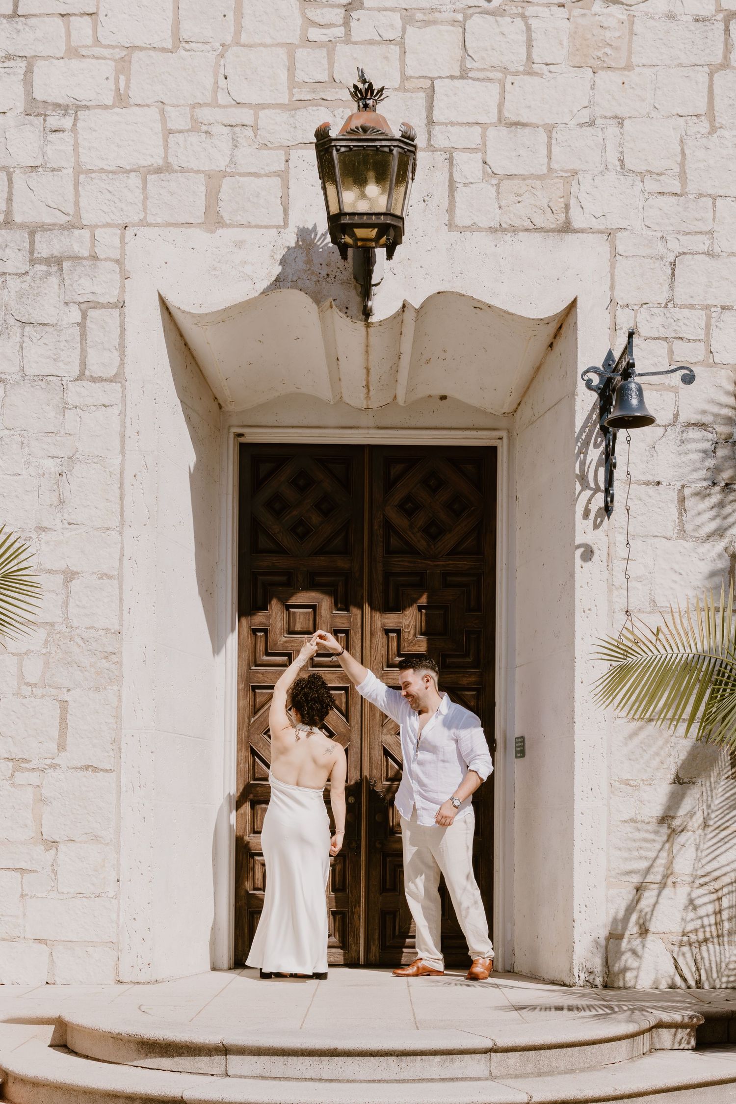 A couple in light colored attire poses playfully in front of an ornate wooden doorway with stone walls.