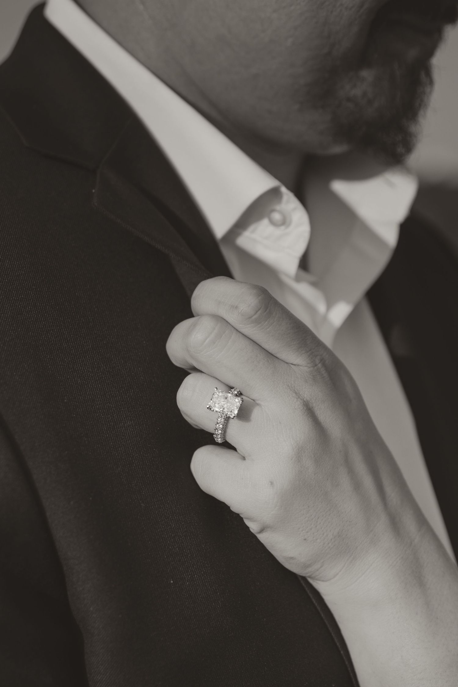 Hand wearing an elegant diamond engagement ring sparkles with light, seen against a dark formal attire.