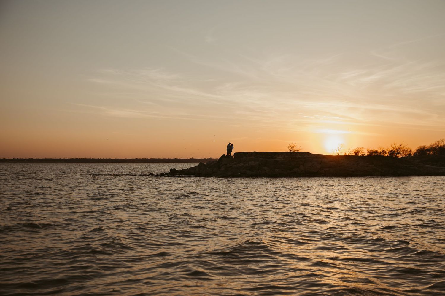 Silhouette of a person standing on rocky shoreline during a golden sunset over calm water.
