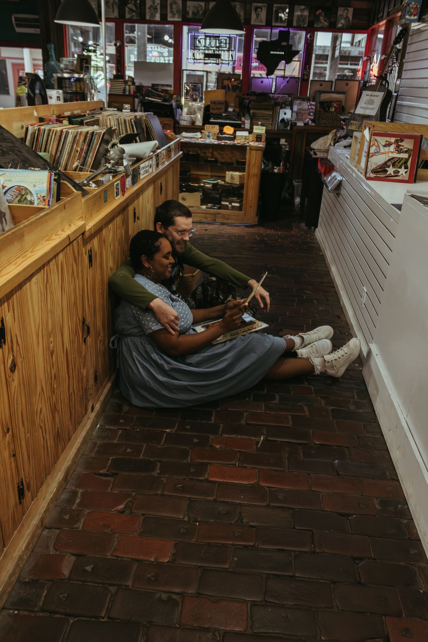 Two people sit on a brick floor browsing vinyl records in a vintage record shop with wooden bins and display cases.