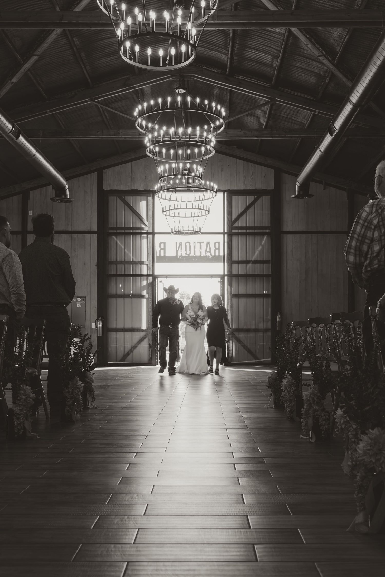 A dramatic black and white view of a barn venue interior with hanging chandeliers and silhouetted figures. Wedding photographer DFW