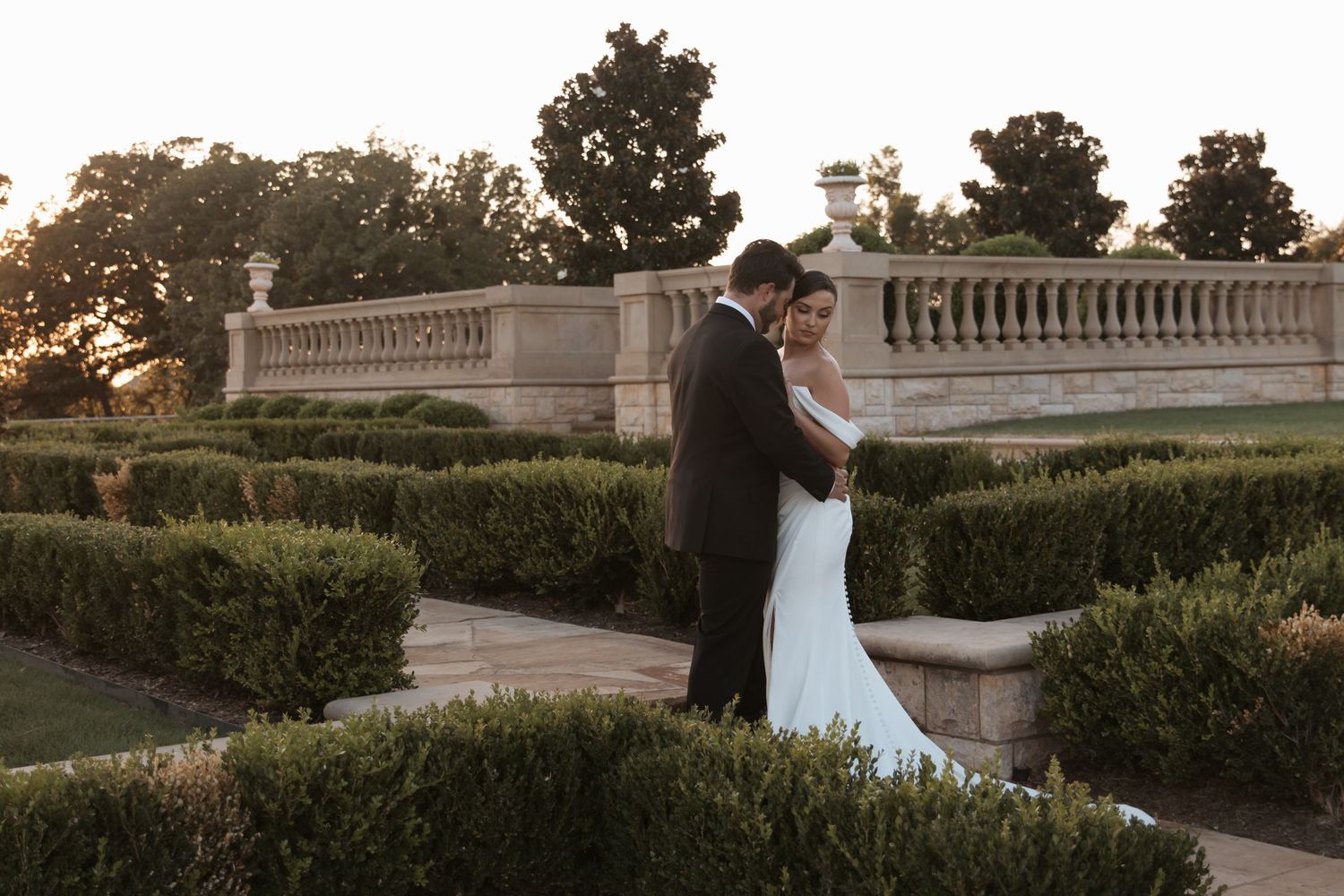 A stone balustrade and manicured hedges create an elegant garden setting at sunset.