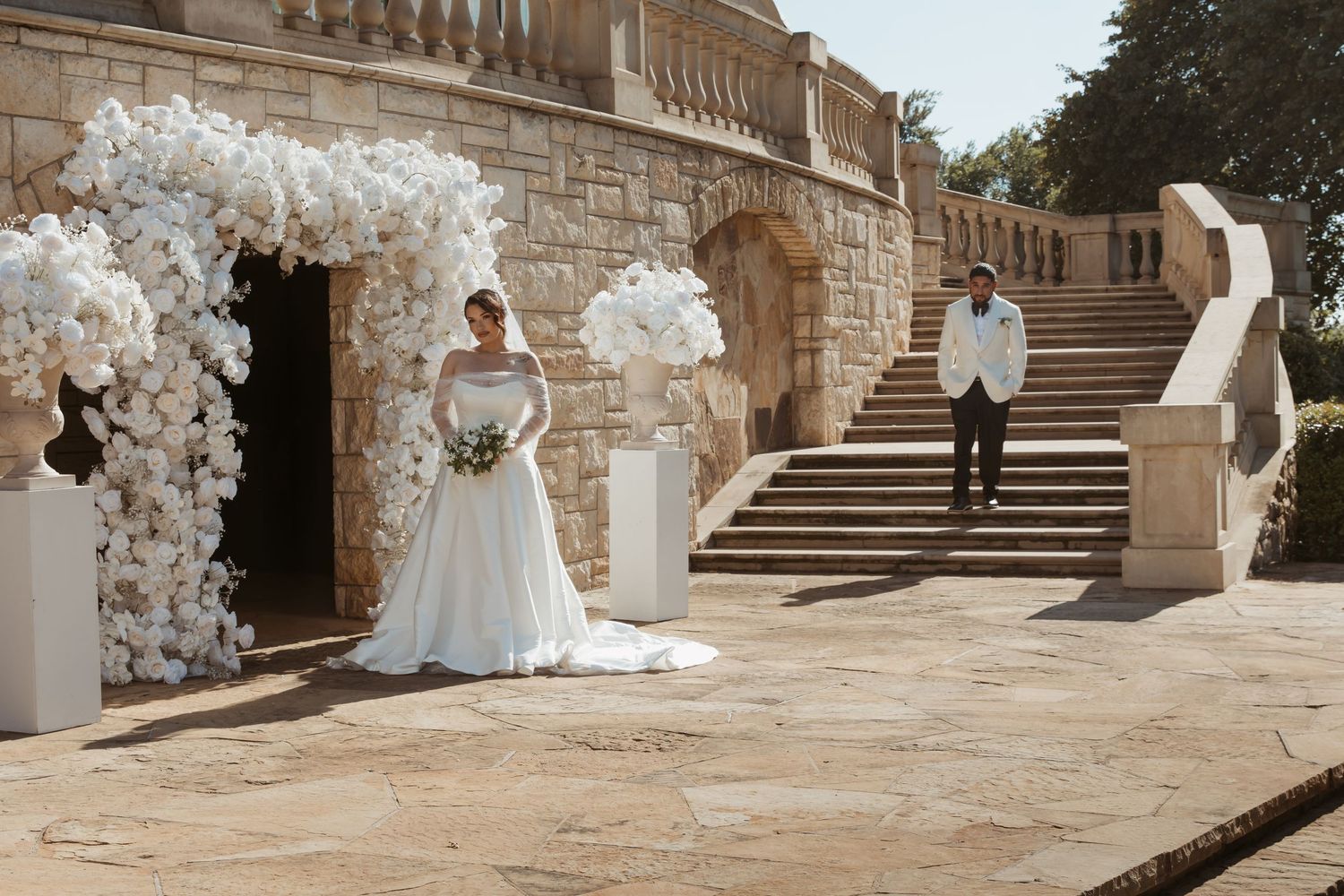 A bride in a white wedding gown poses in front of a stone staircase decorated with elegant white floral arrangements.